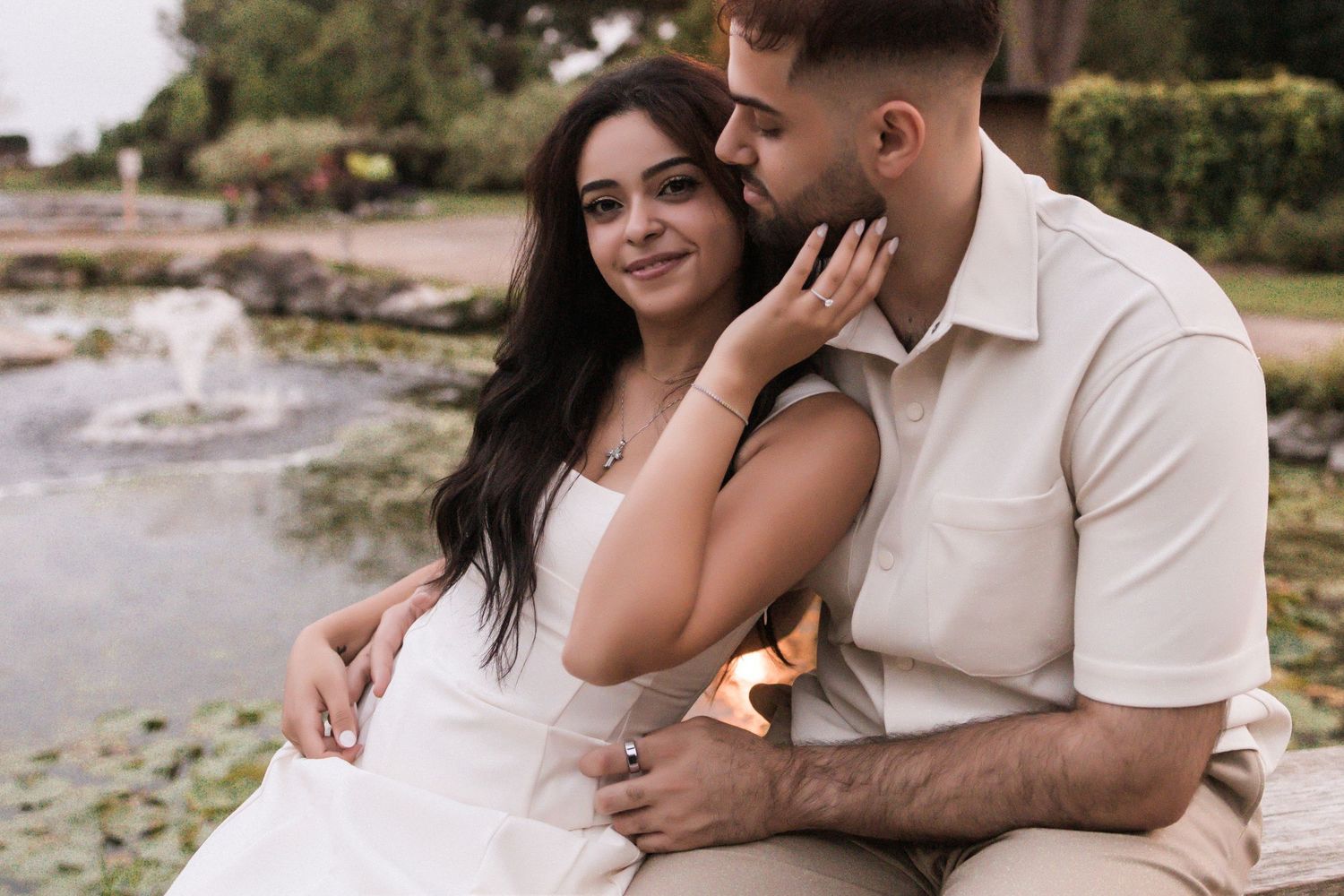 A romantic couple embraces by a peaceful waterfront on a sunny afternoon wearing light colored clothes.