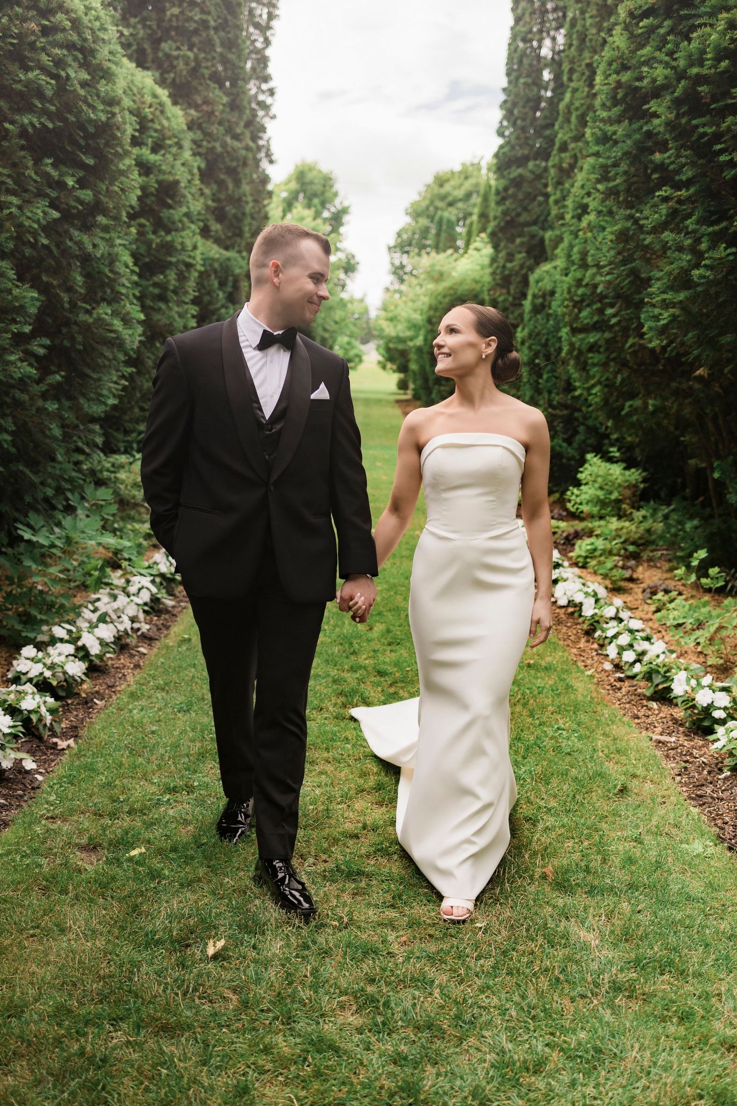 A couple walks together along a garden path lined with white flowers and evergreen trees.