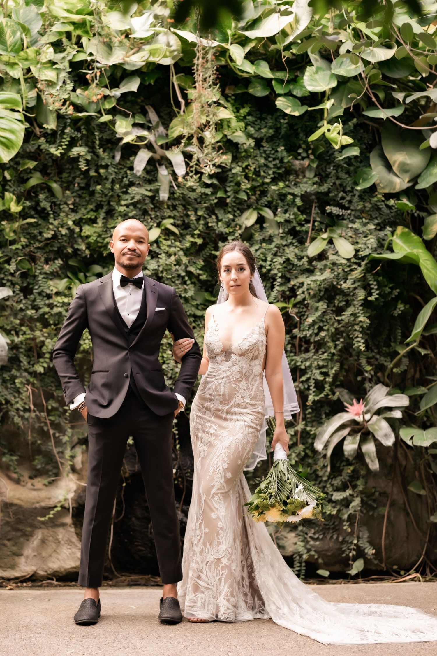 A couple poses against a lush garden wall background in formal wedding attire with a long trailing bridal gown.