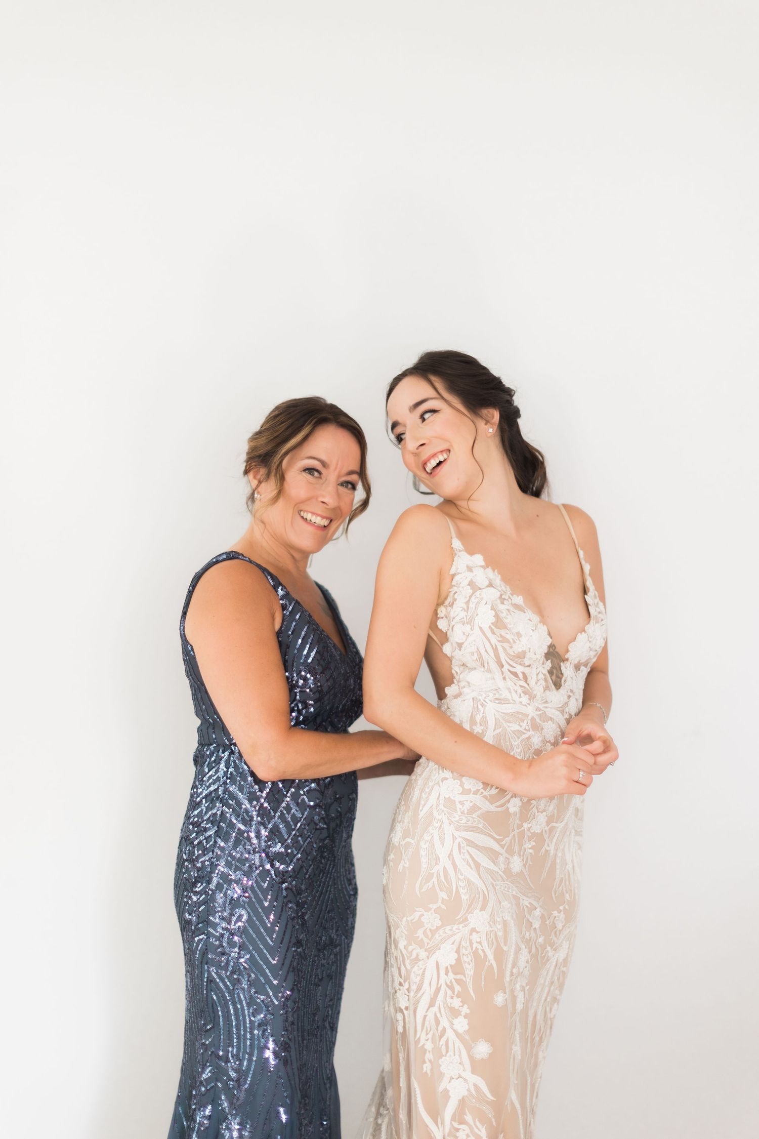 Two women laugh together while wearing formal dresses against a white background.