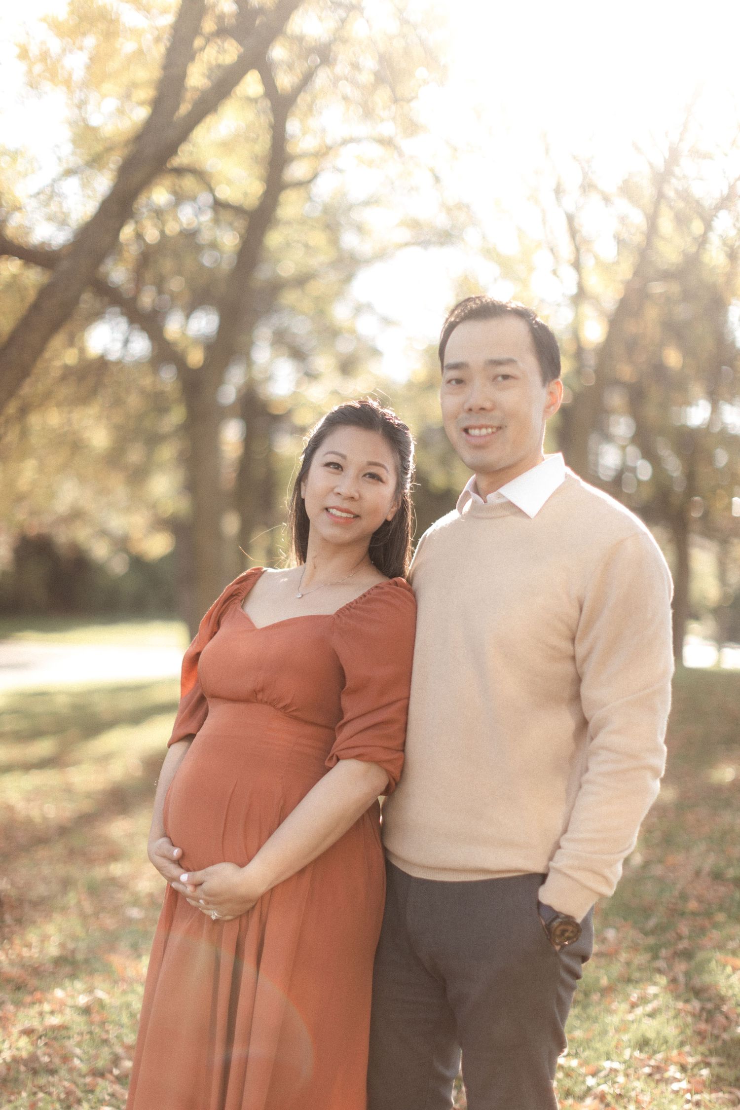 A couple poses for a maternity photo in a park with warm autumn light filtering through the trees.
