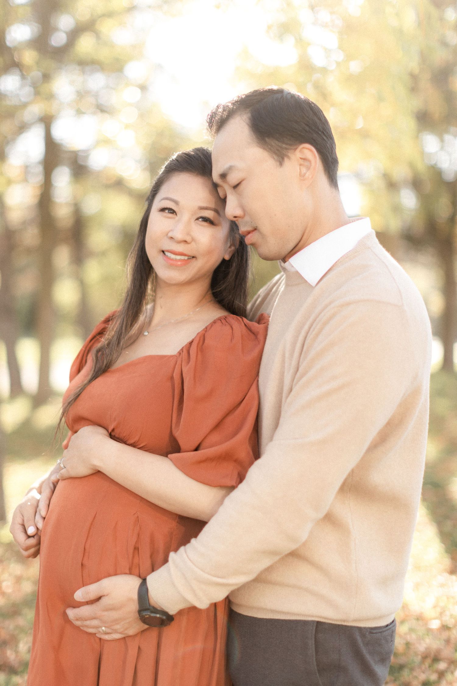 Couple poses together in autumn forest setting with woman in rust-colored dress and man in beige sweater.