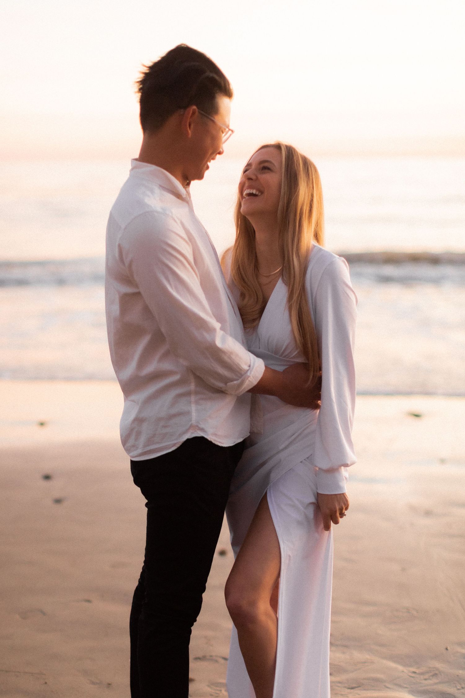 Romantic couple laughing together during sunset photoshoot at the beach wearing matching white outfits.