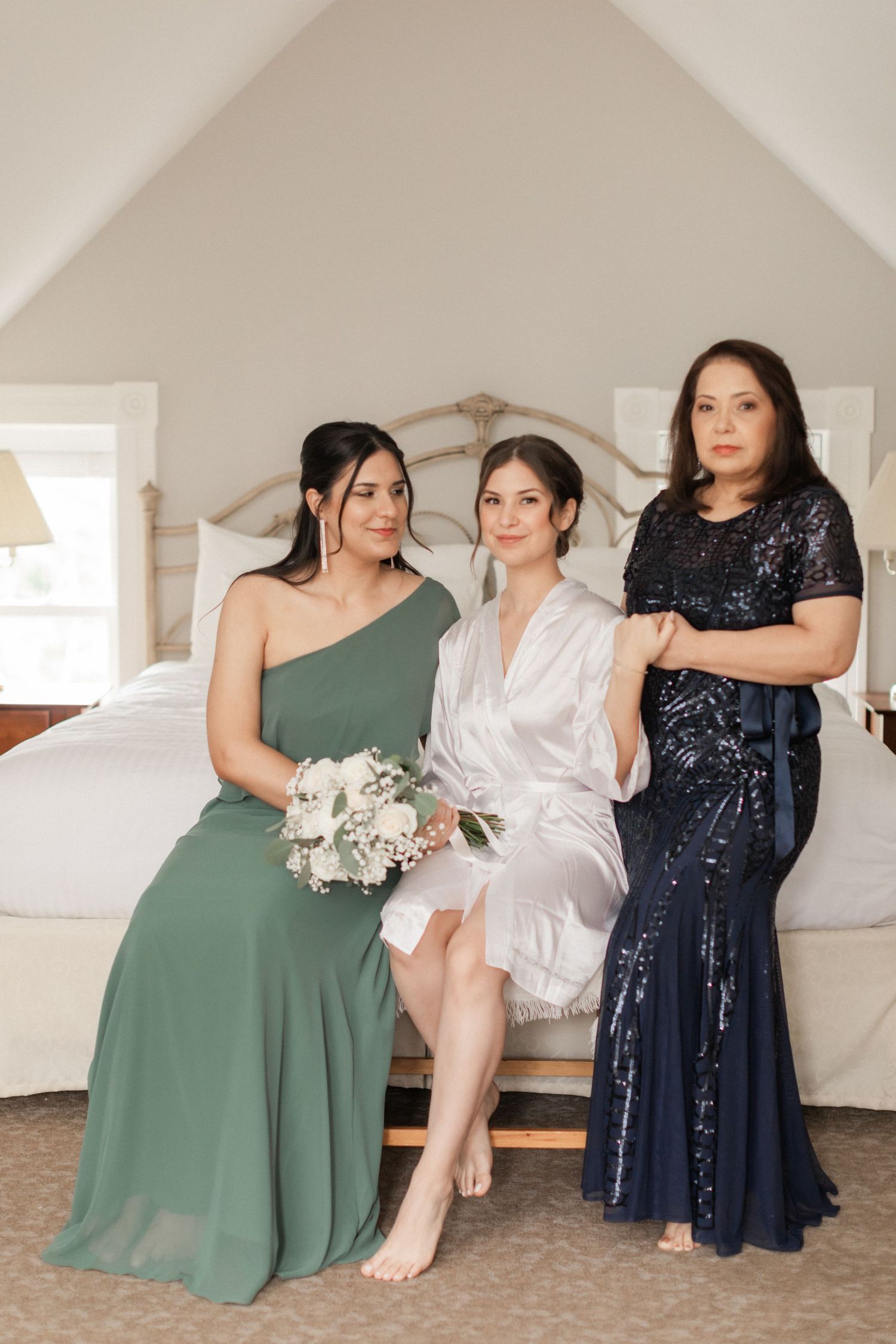 Wedding party in elegant dresses seated on bed in bright bedroom.