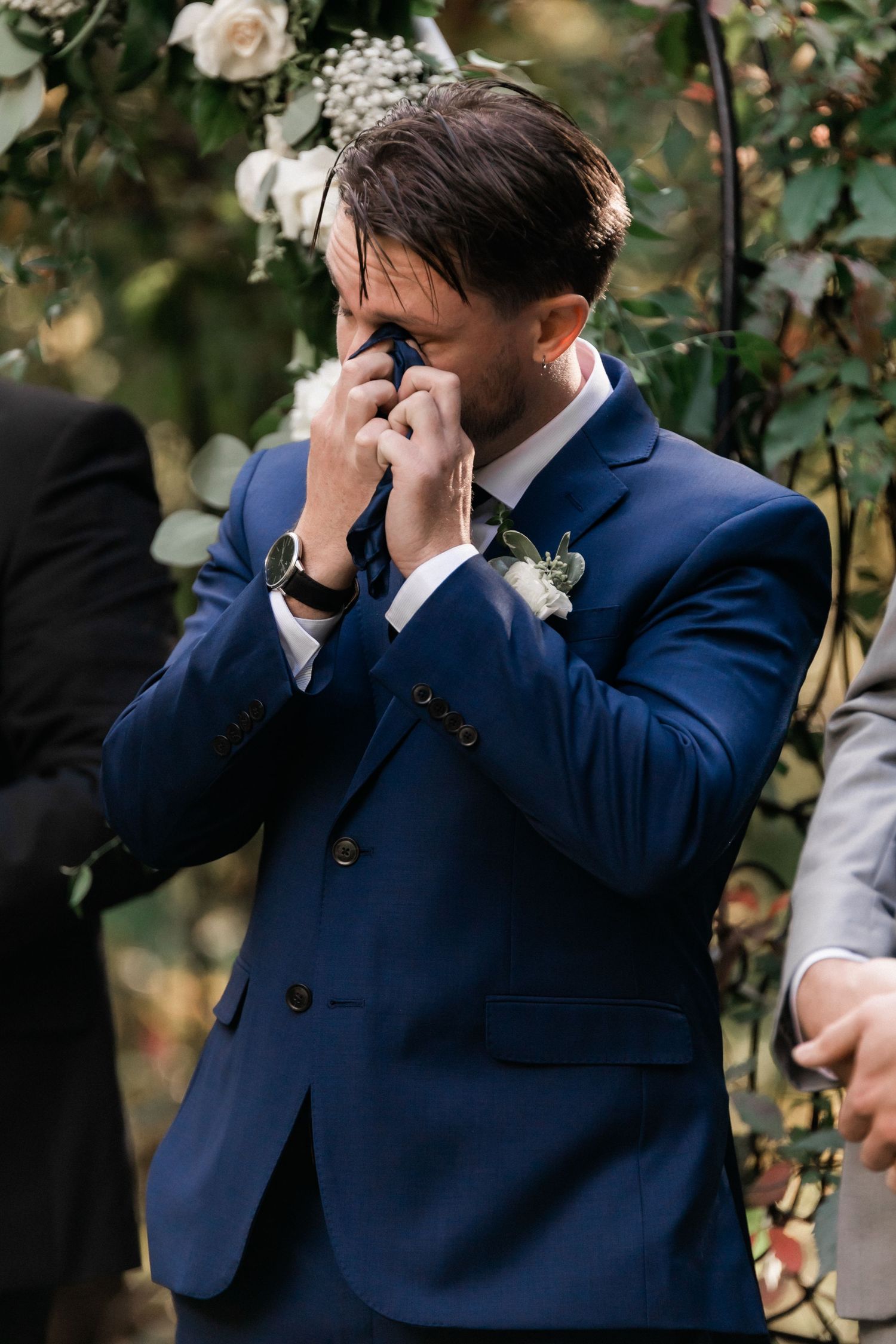 Emotional groom in navy suit wipes tears during outdoor wedding ceremony.