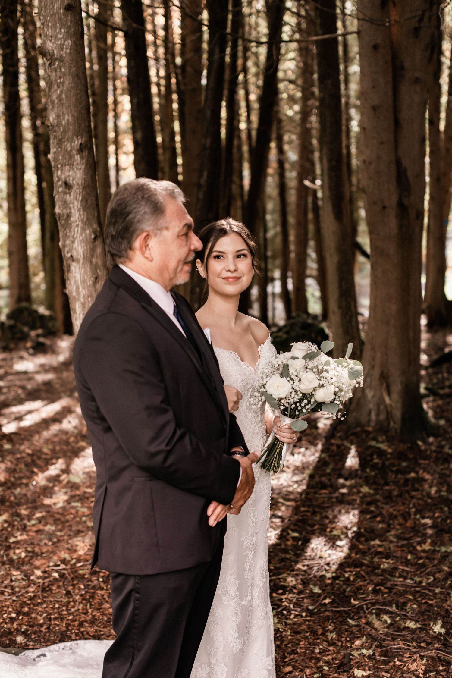 A romantic forest wedding portrait featuring elegant attire and a white floral bouquet amidst tall trees.