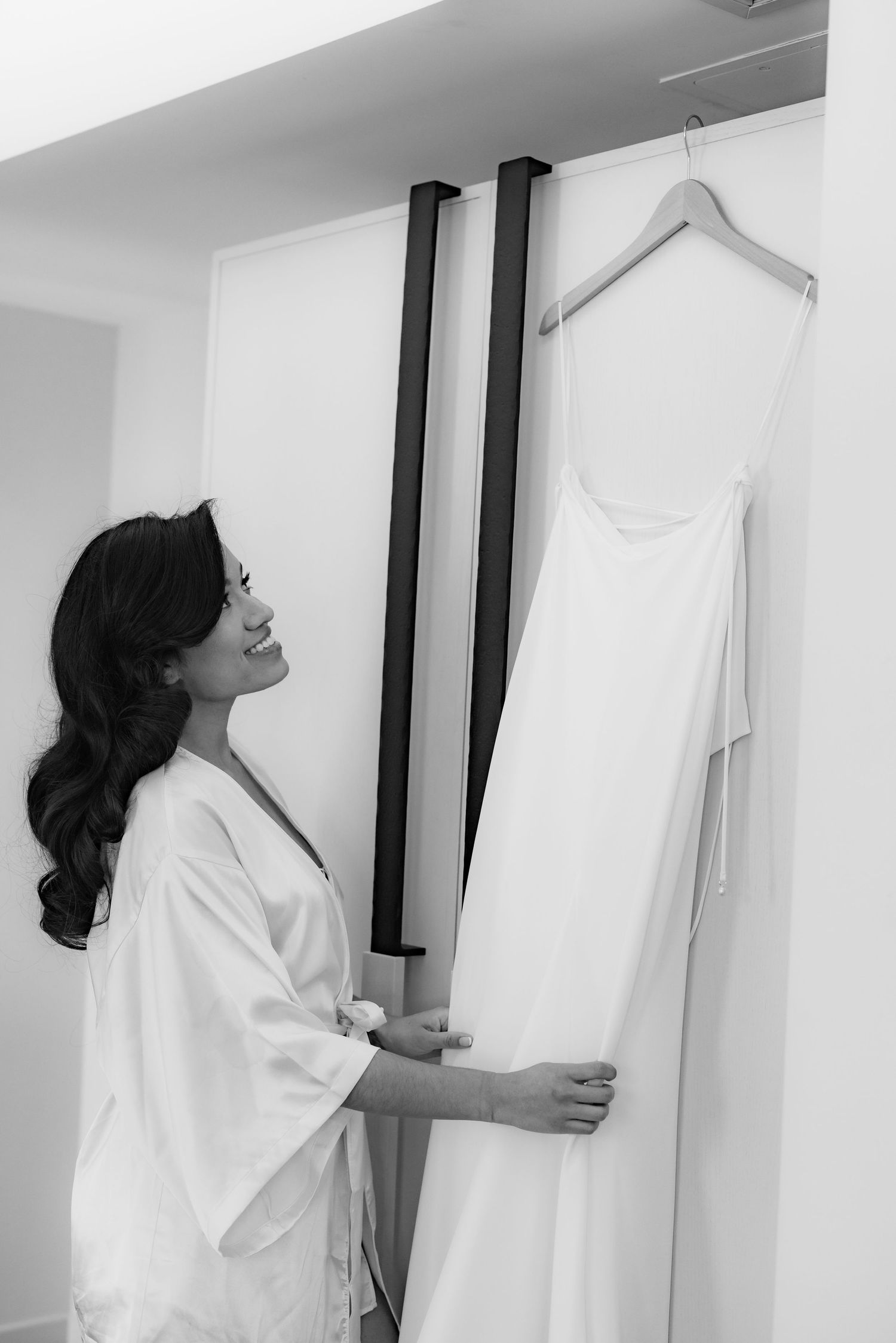 Bride in white robe admiring wedding dress hanging on display.
