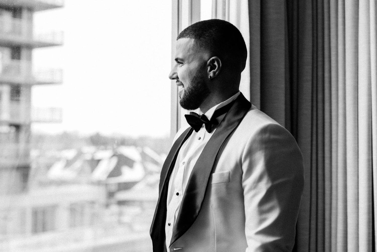 A groom in formal attire gazes out of a window in black and white.