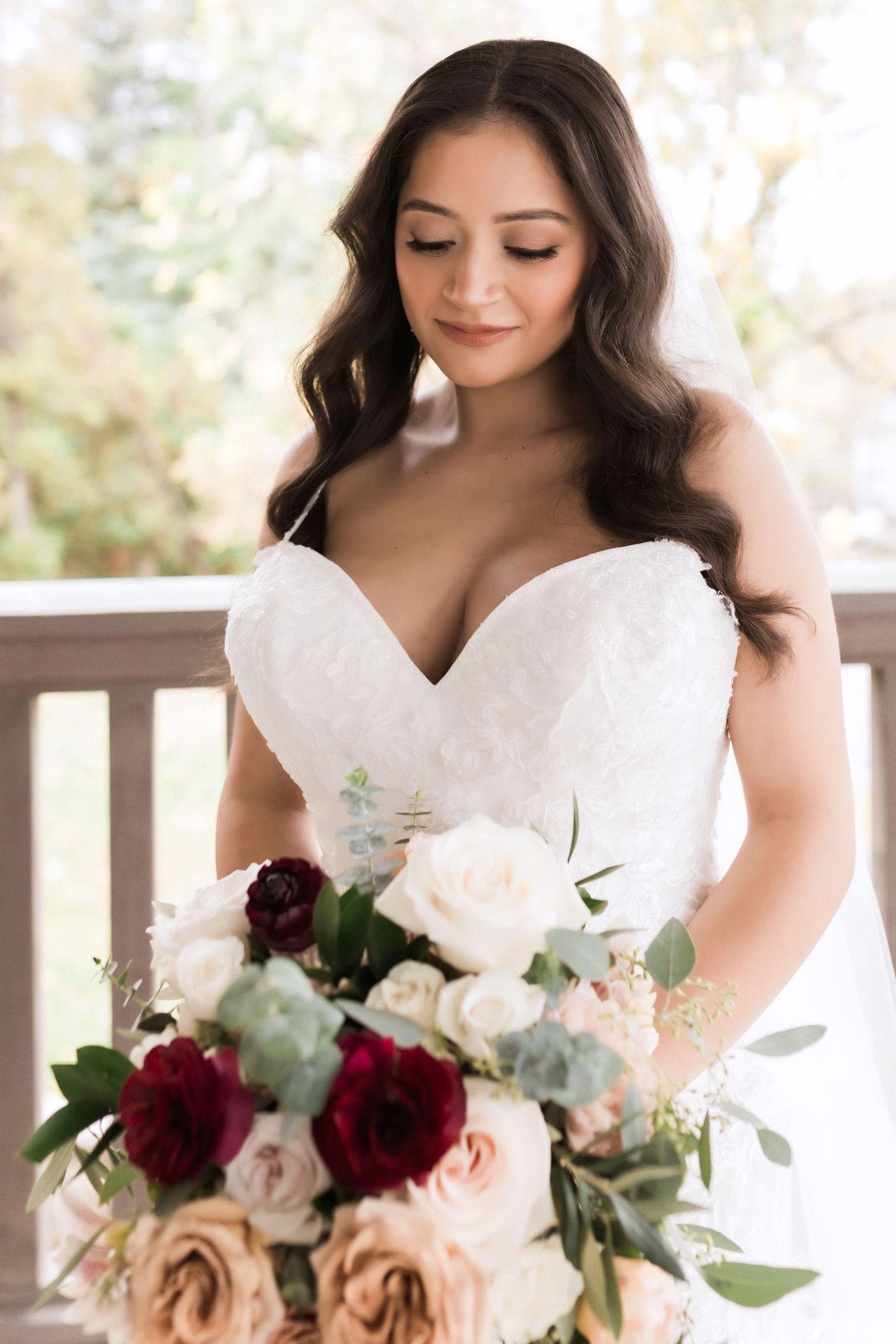 A bridal bouquet features deep burgundy and blush roses with eucalyptus greenery against a white dress.