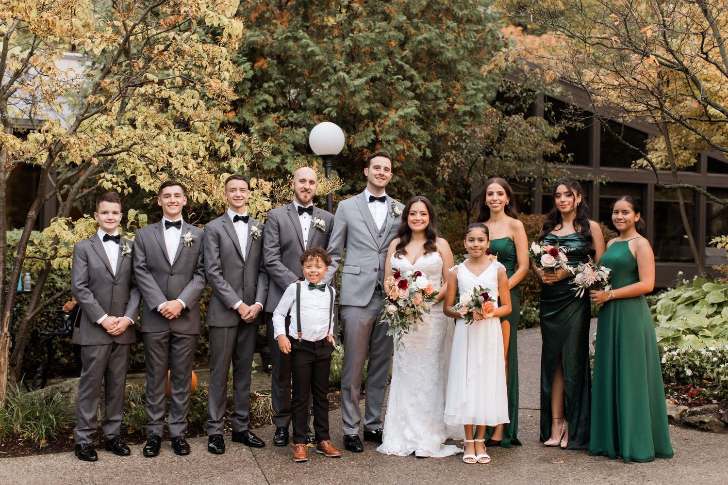 Wedding party poses outdoors with groomsmen in grey suits and bridesmaids in green dresses.