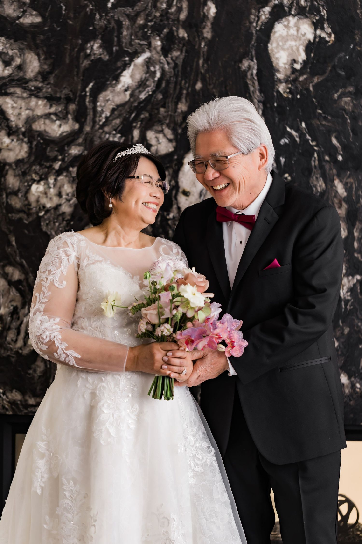 Joyful mature couple in wedding attire sharing laughter against marble backdrop.