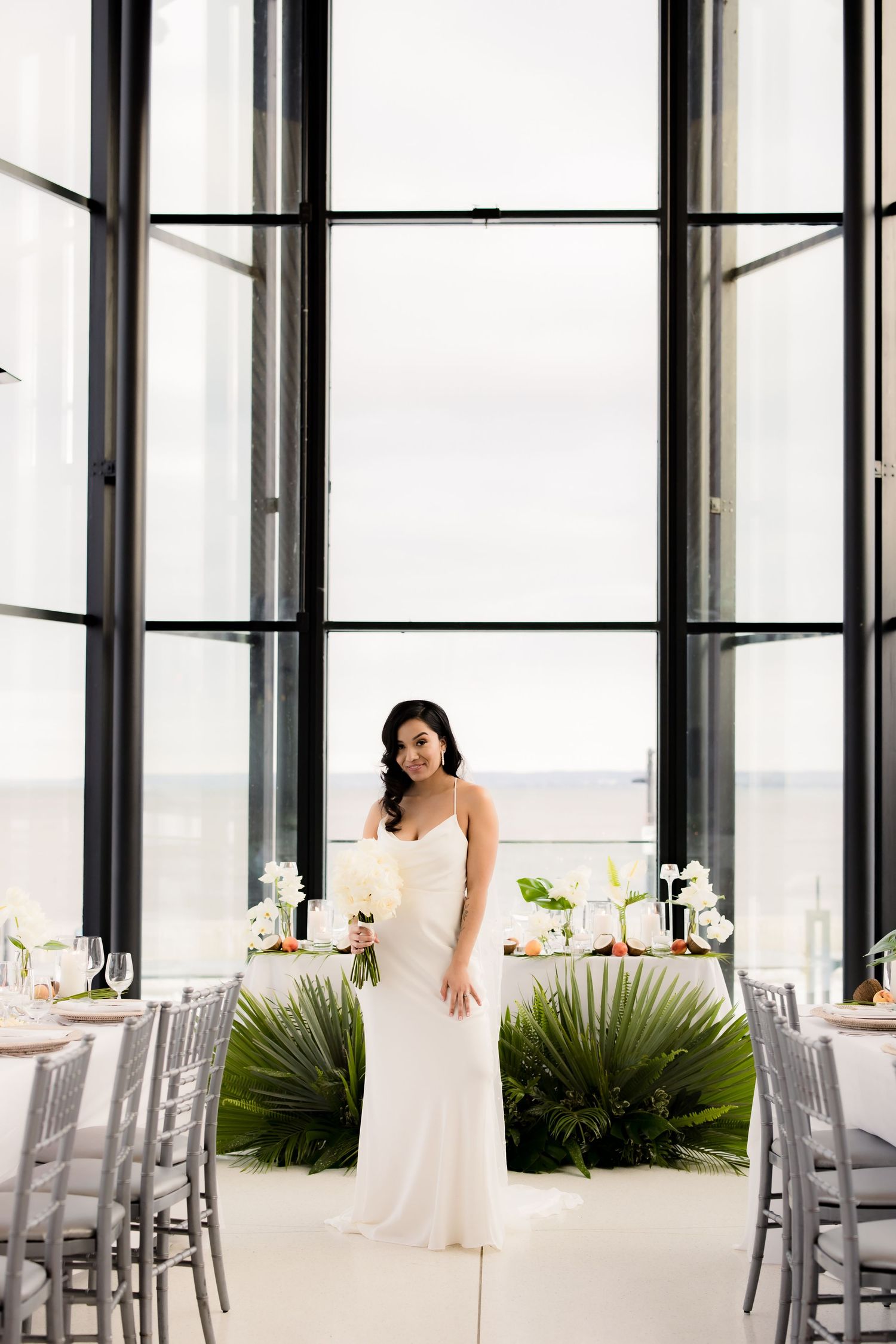 Elegant bride in white gown poses in modern venue with floor-to-ceiling windows and silver decor.