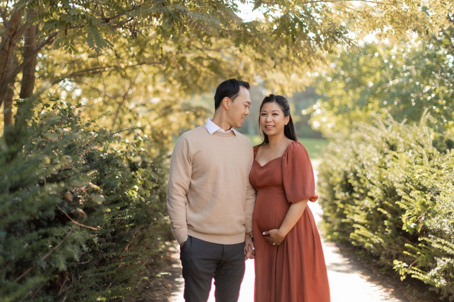 Expectant parents walking along tree-lined path during golden hour maternity session.