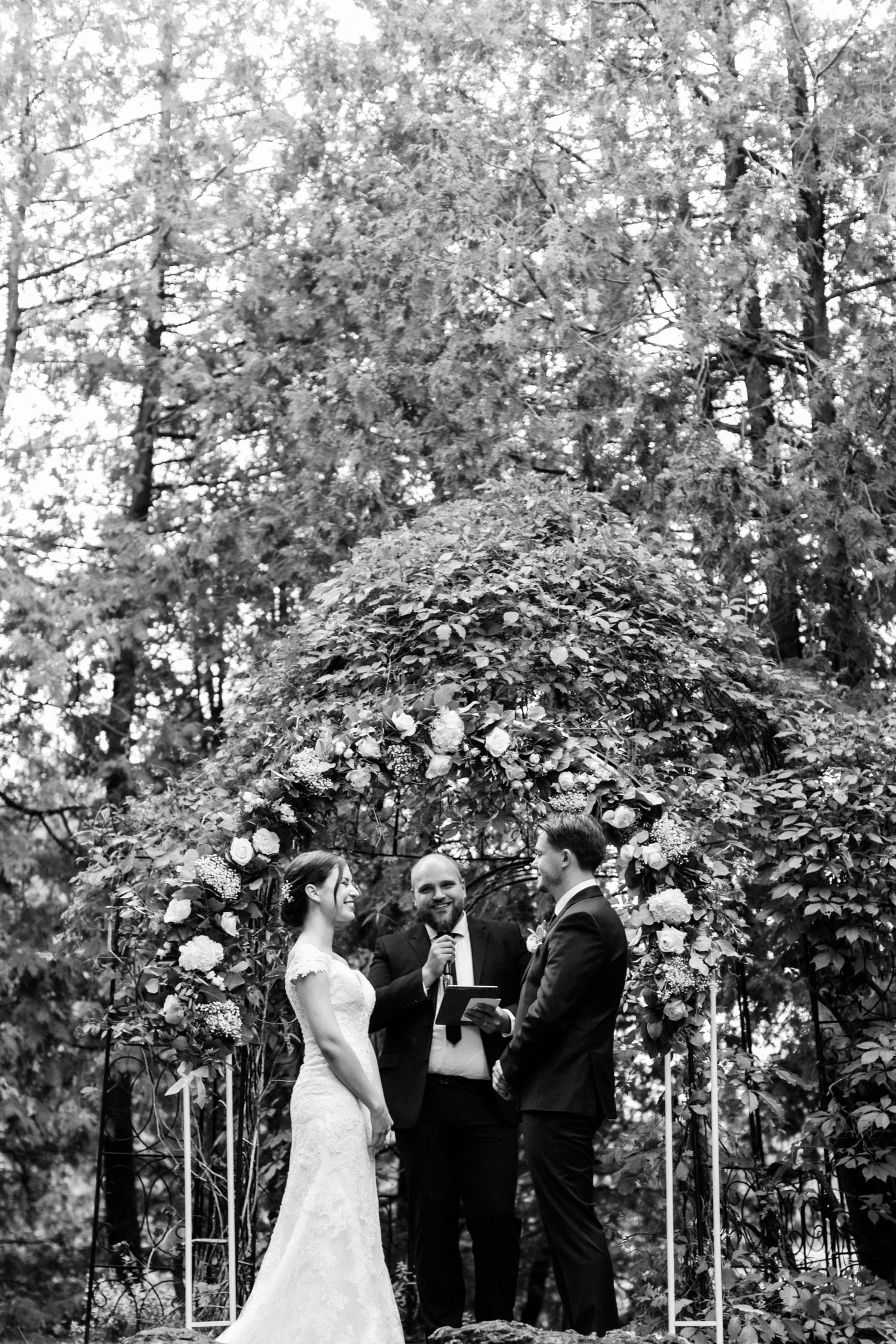 Black and white photo of a wedding ceremony under a floral arch in a forest setting.