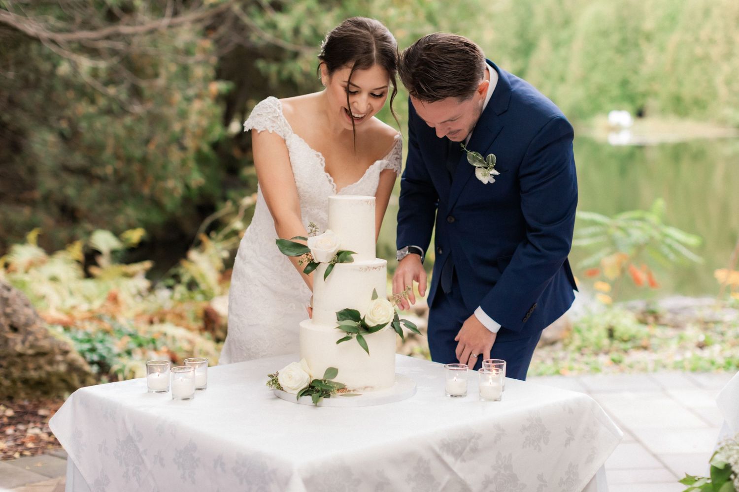 Couple cutting their white wedding cake together at outdoor reception by lakeside.