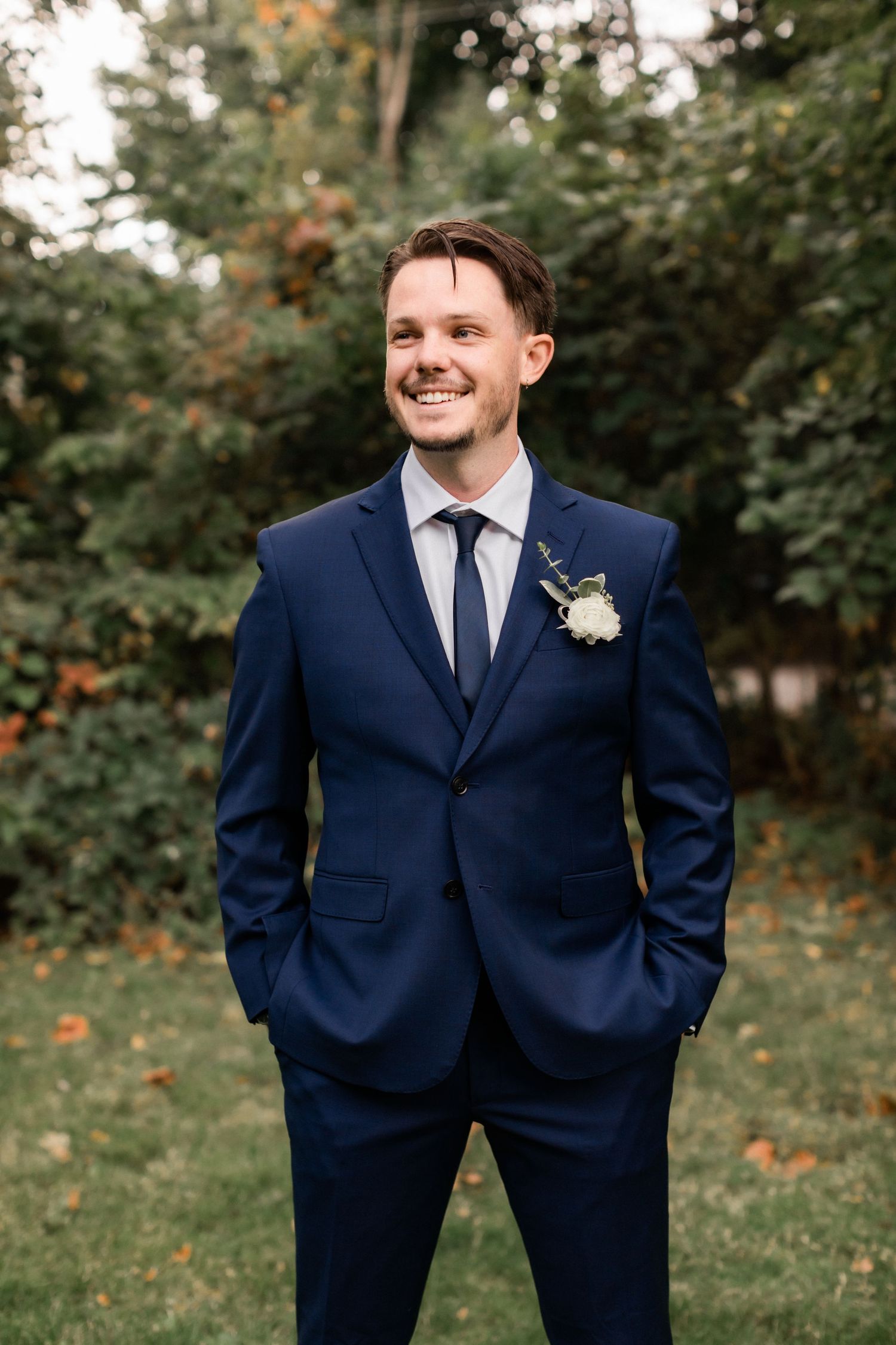 Groom in navy suit with boutonniere smiling outdoors against green foliage background.