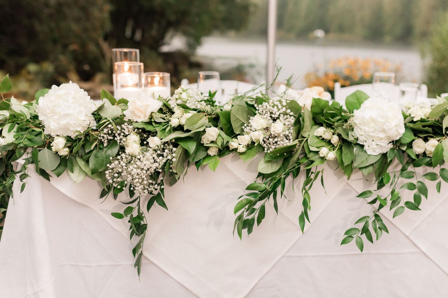 Elegant white floral table decorations with greenery garlands at an outdoor wedding reception.
