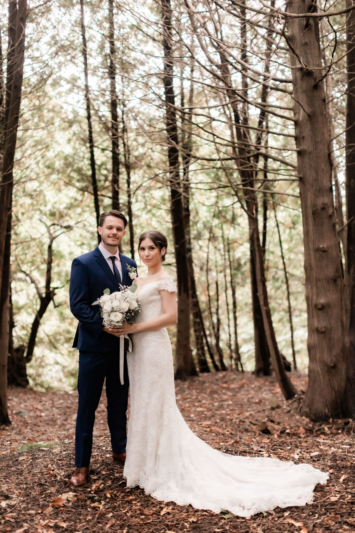Romantic forest wedding portrait with couple standing among tall trees in autumn setting.