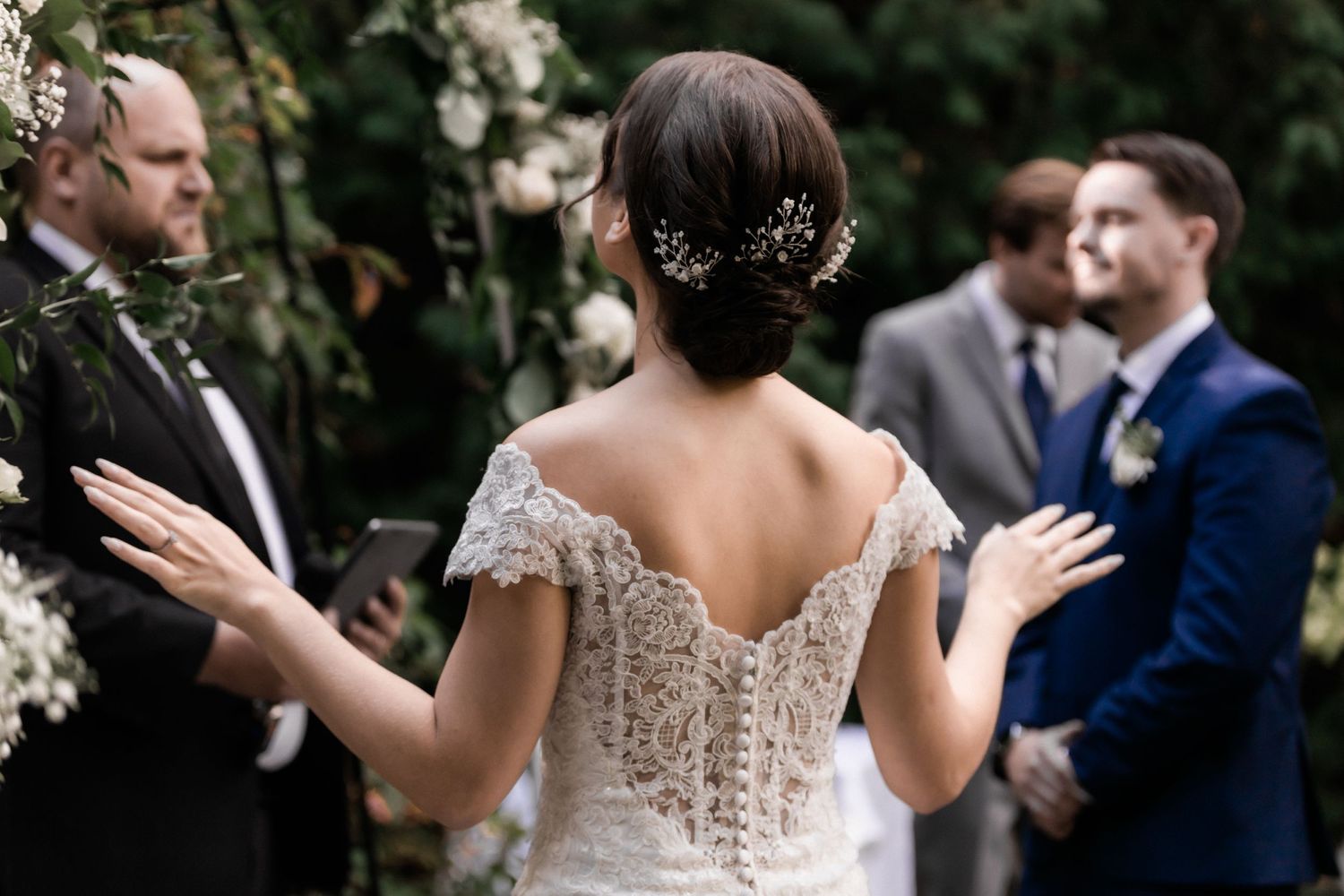 A bride in a lace wedding dress with delicate back detail gestures during an outdoor ceremony.