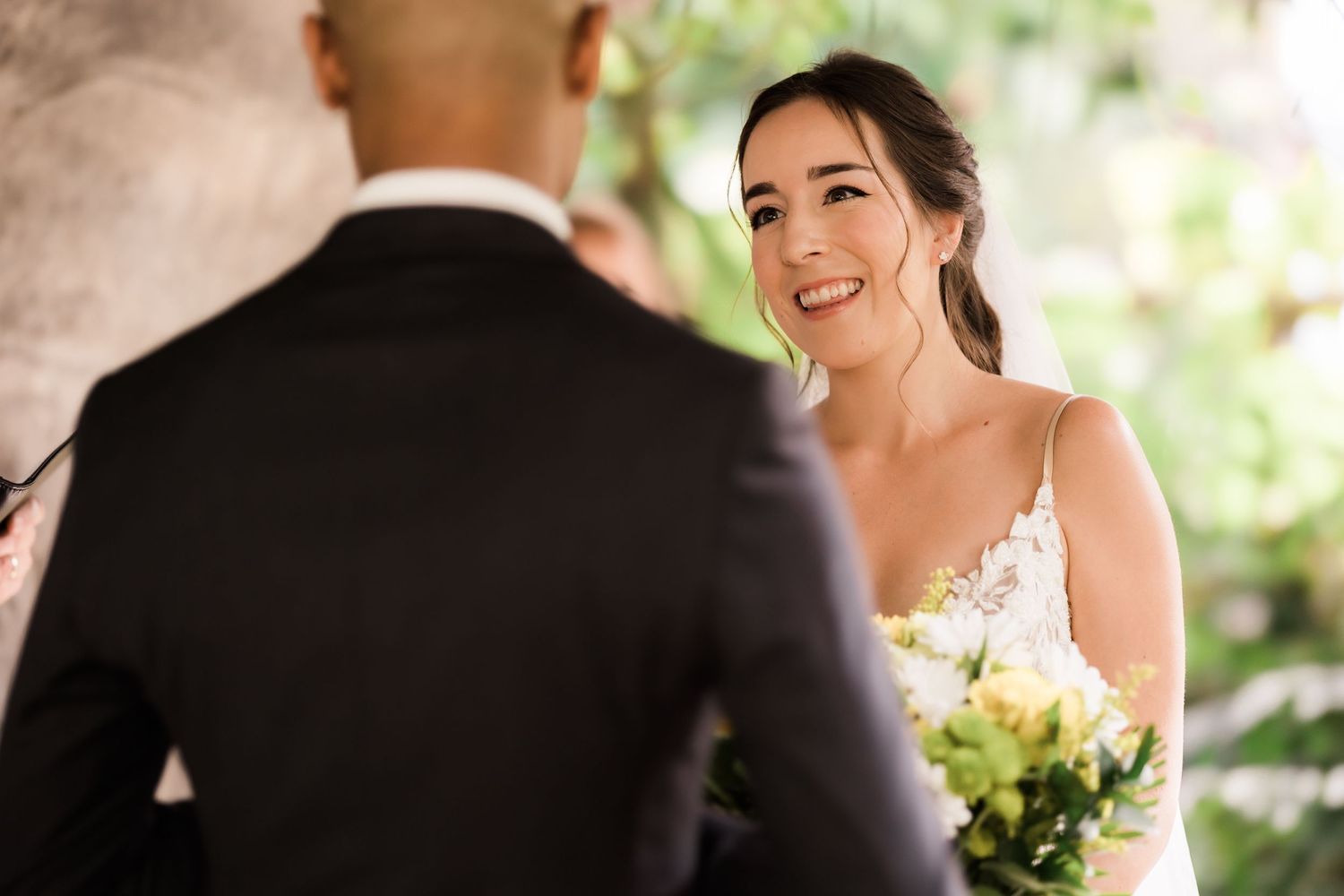 Bride smiles joyfully during outdoor wedding ceremony surrounded by greenery.