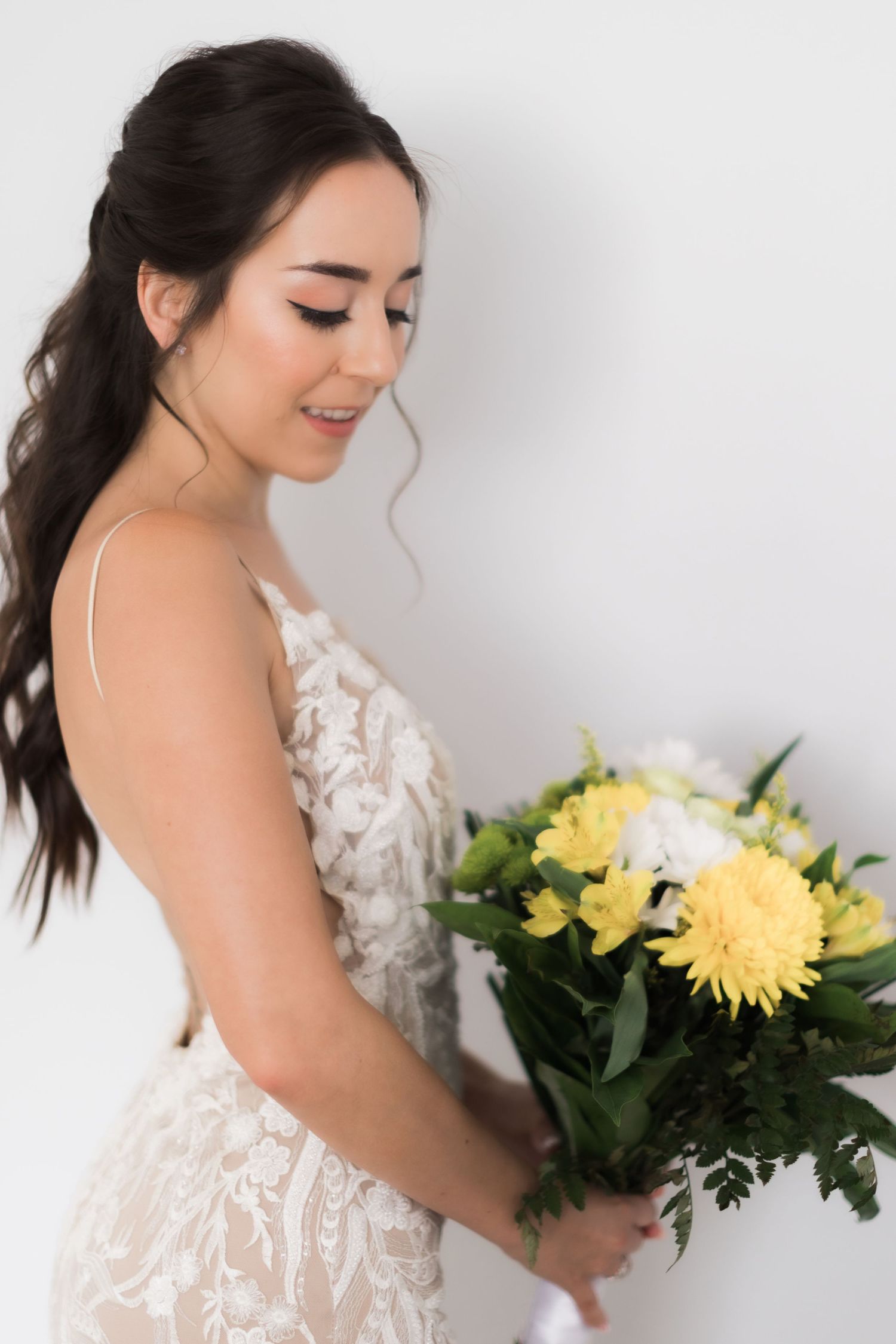 Bride wearing delicate lace wedding gown smiles while holding yellow and white floral bouquet.