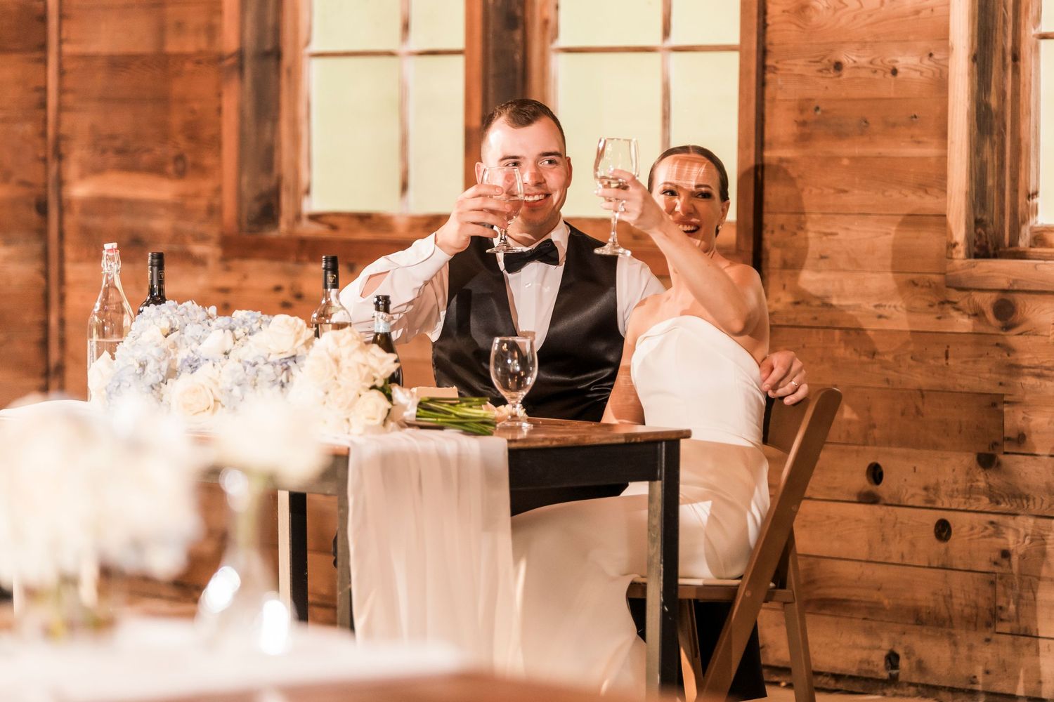 Reception guests seated at long tables in a rustic barn venue with natural window light.