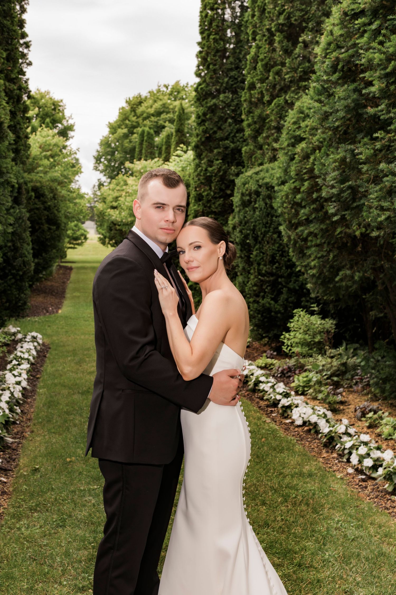 Elegant wedding couple poses in formal attire along a manicured garden path with tall hedges.