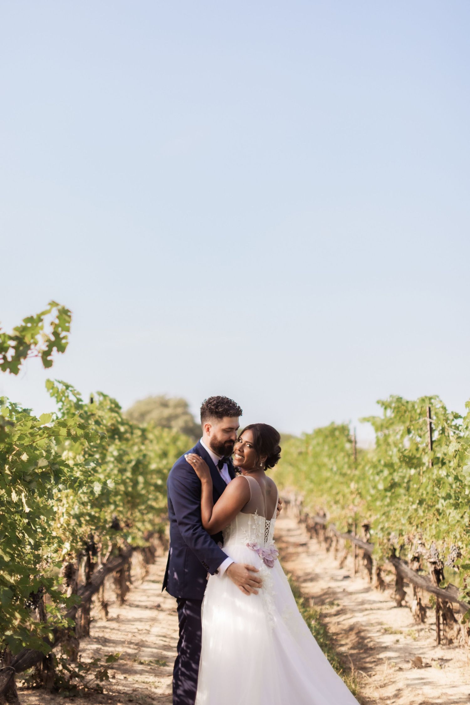Romantic wedding couple shares intimate moments while strolling through sun-drenched vineyard rows on their special day.