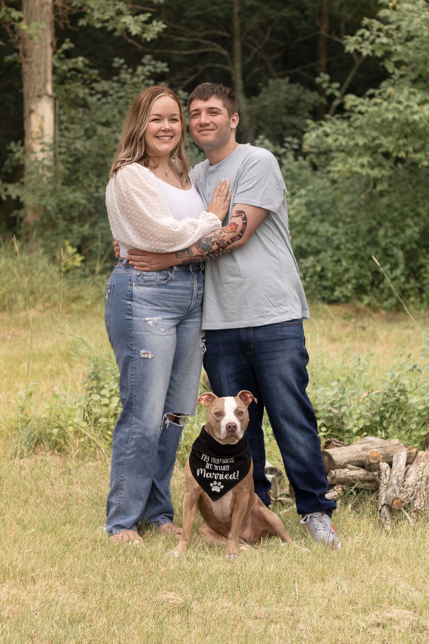 Happy couple poses with their brown dog wearing a bandana in a grassy outdoor setting.
