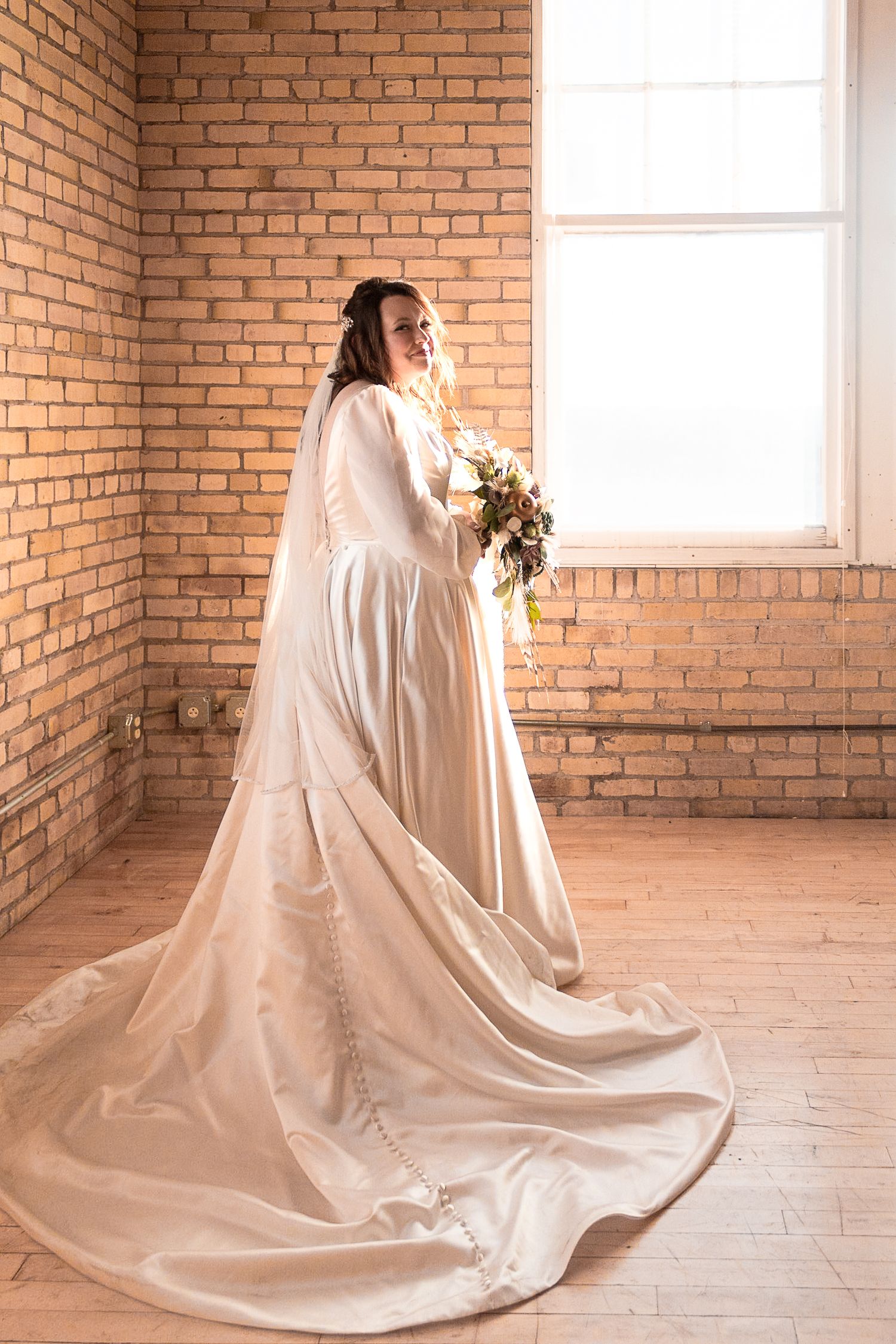 Bride in flowing white wedding dress poses beside brick wall with natural window light.