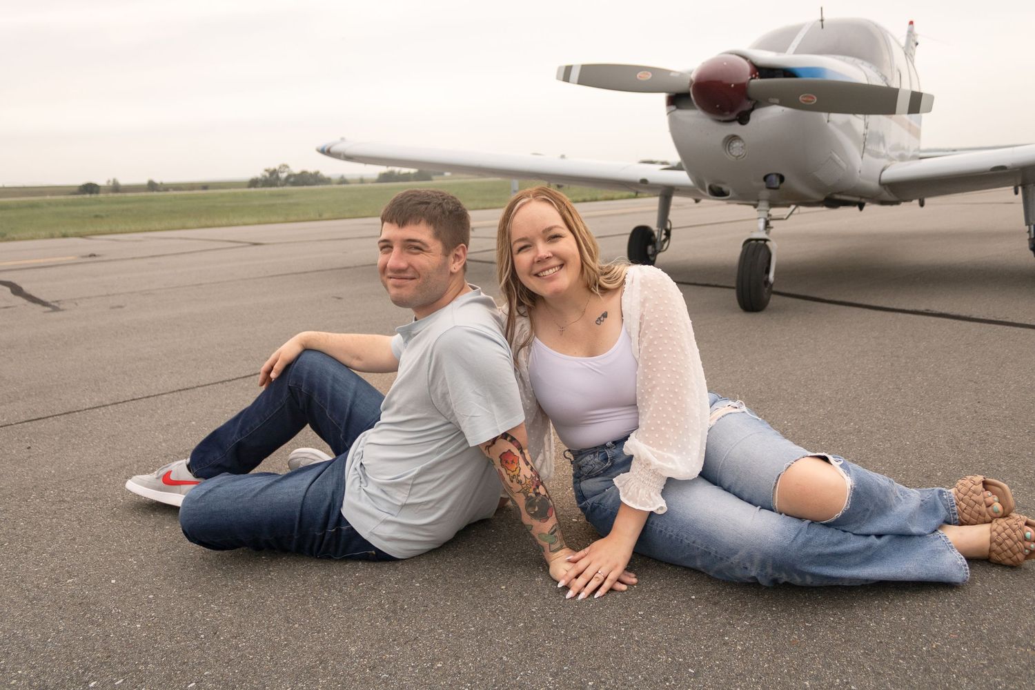 Couple sitting back-to-back on runway with small aircraft in background.