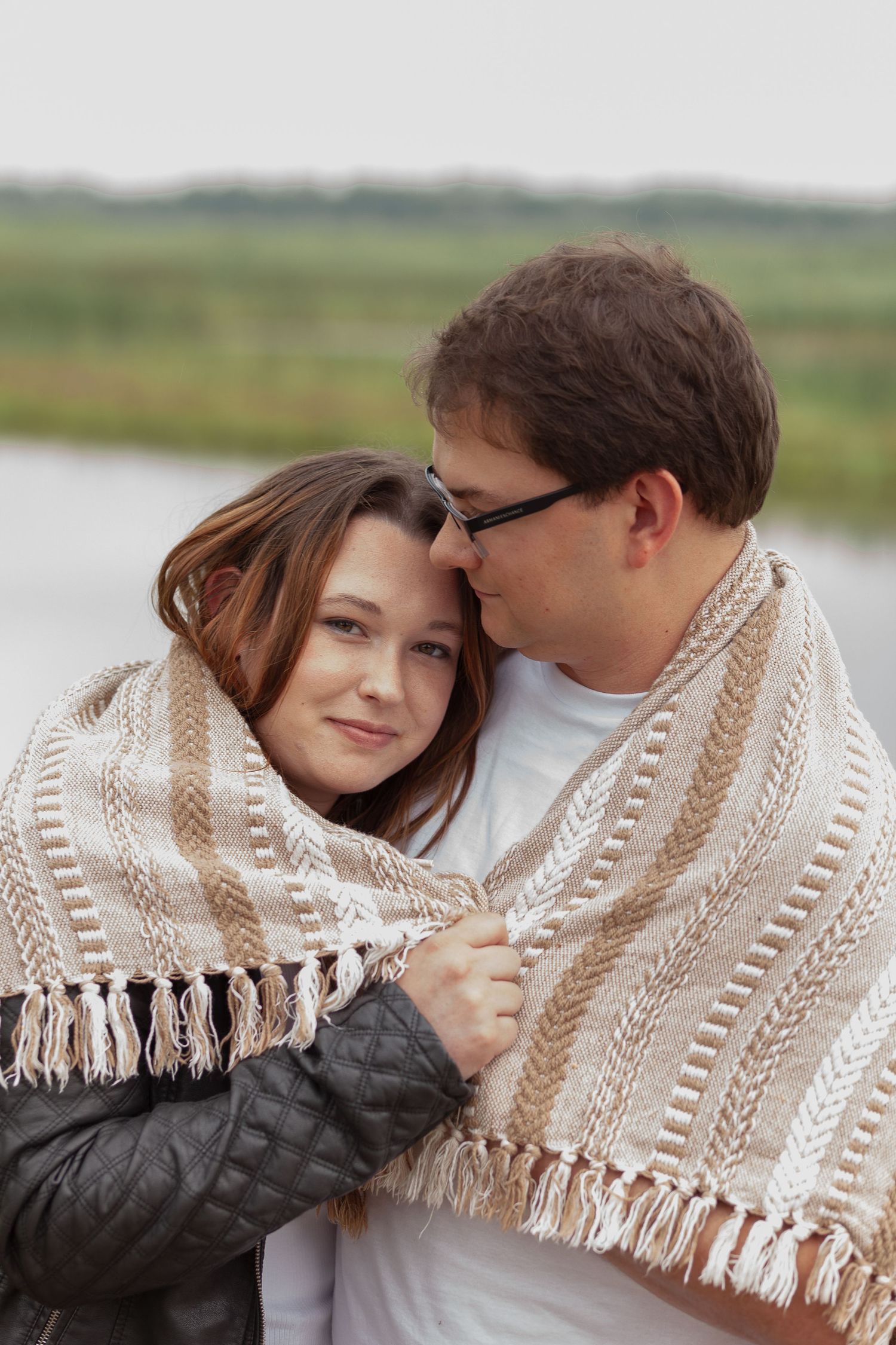 Intimate couple wrapped in knit blanket share tender moment by waterfront.