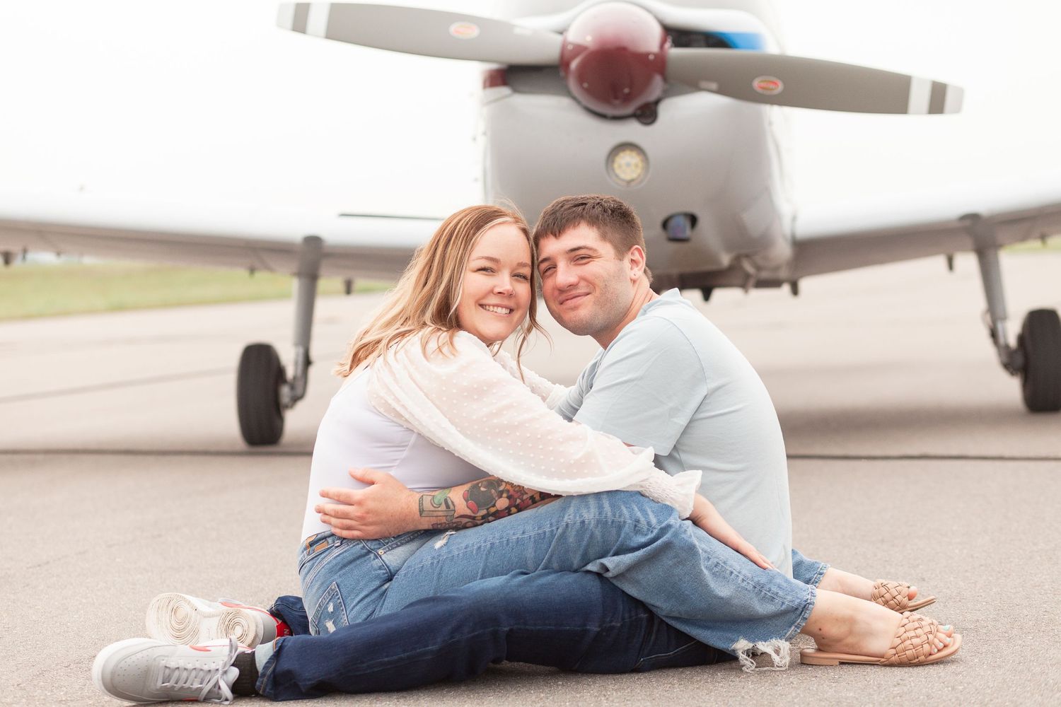 A couple sits together on a runway with a small aircraft in the background.