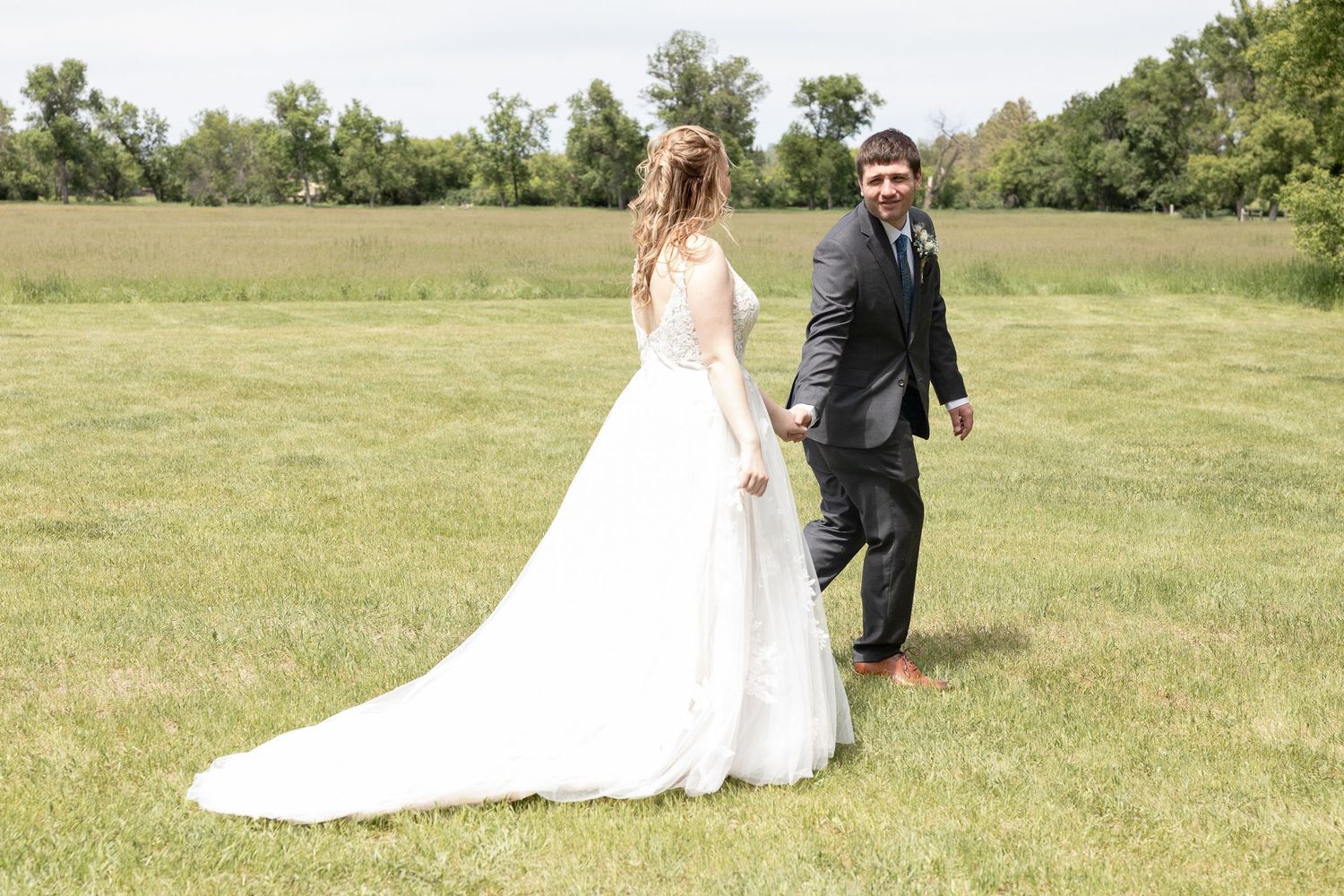 Couple walking through open field in wedding attire with long white train trailing behind.