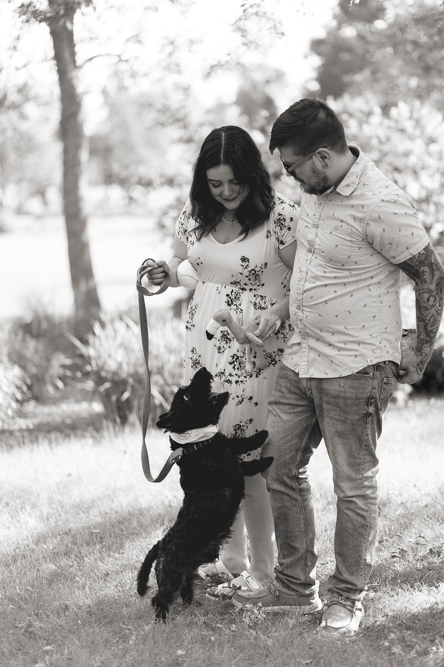 Black and white photo of couple playing with energetic dog outdoors in park setting.