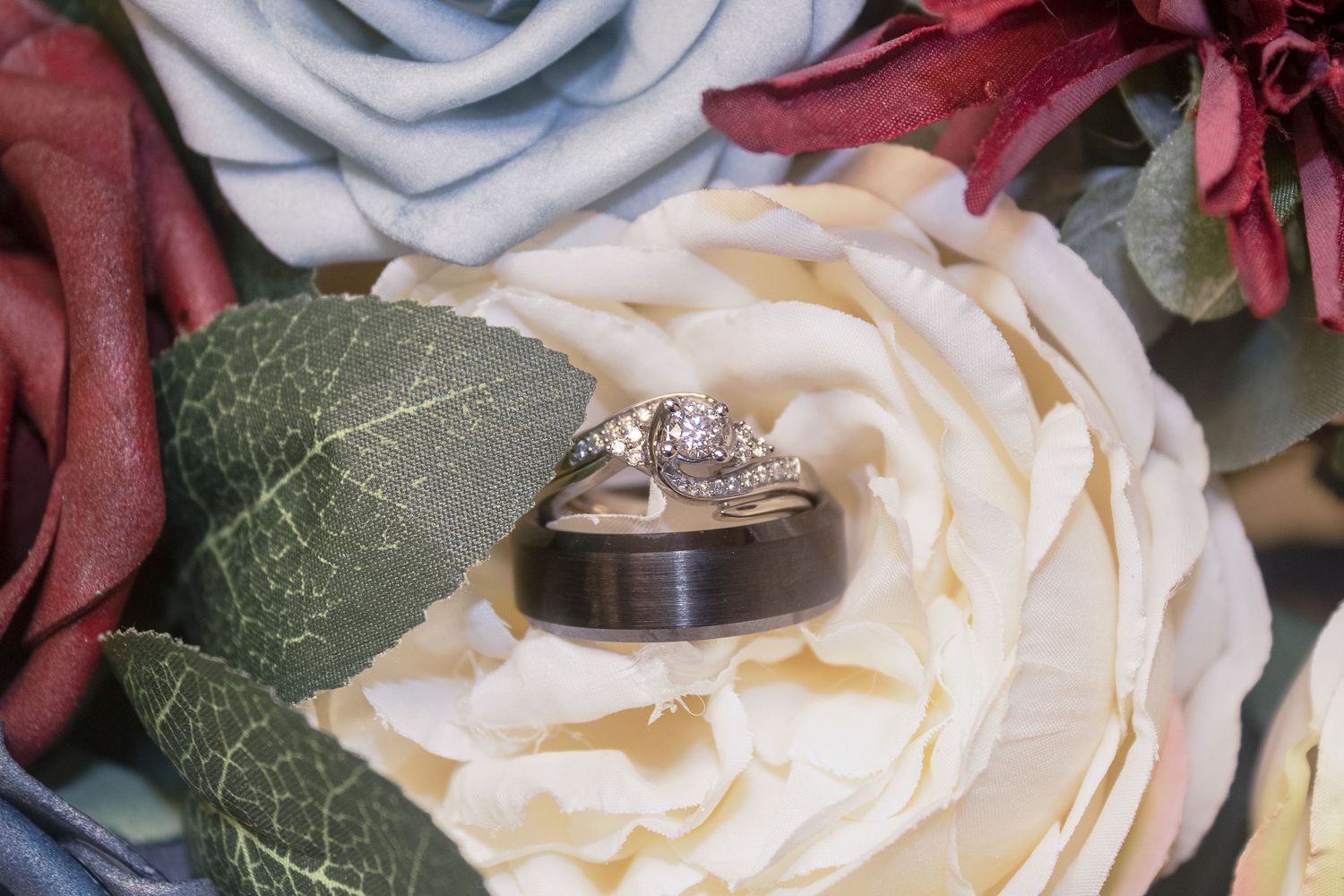 Wedding rings nestled on a cream rose with burgundy floral accents.