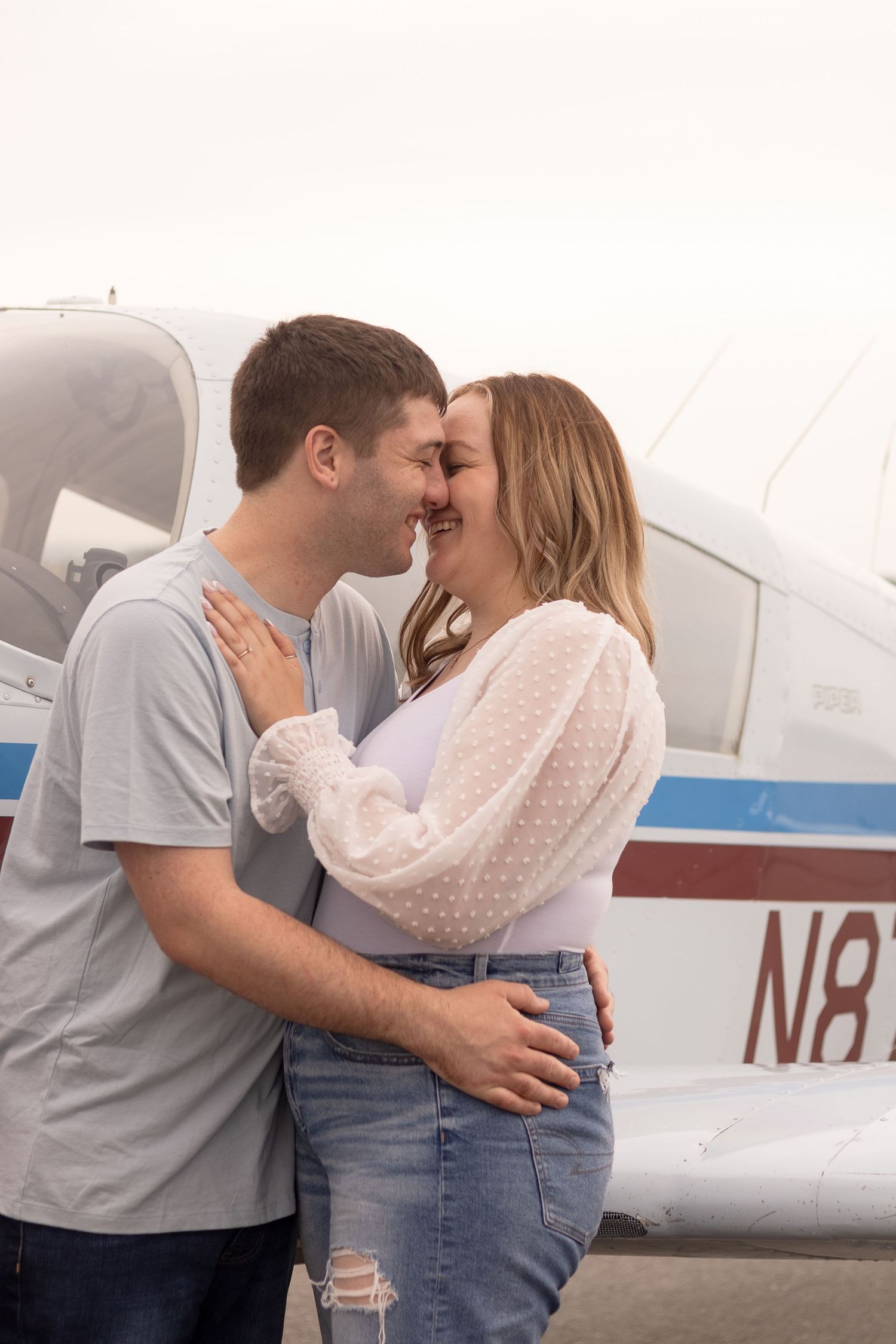 Romantic couple embracing in front of small private airplane during engagement photoshoot.
