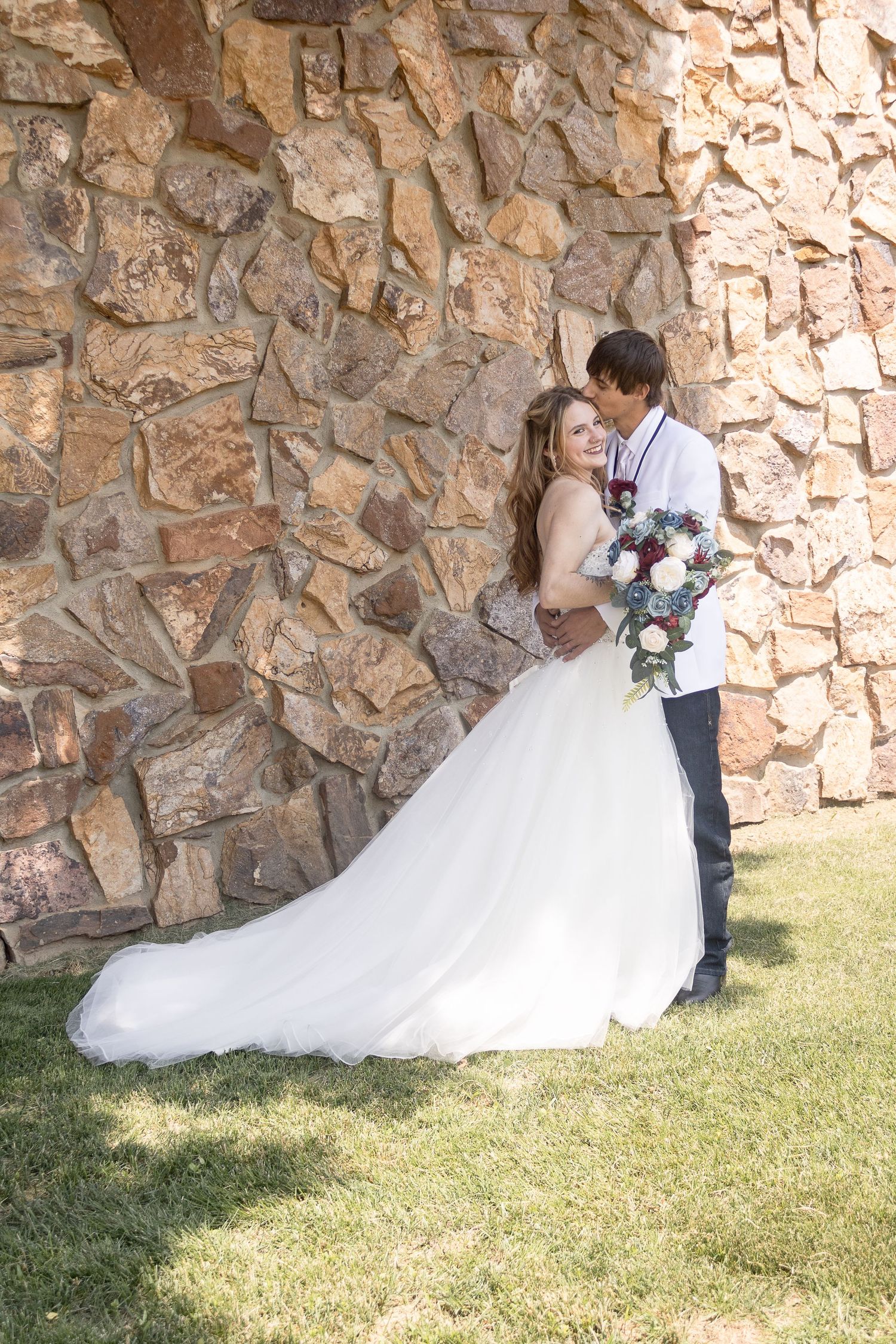 Wedding couple poses against rustic stone wall with flowing white dress and colorful bouquet.