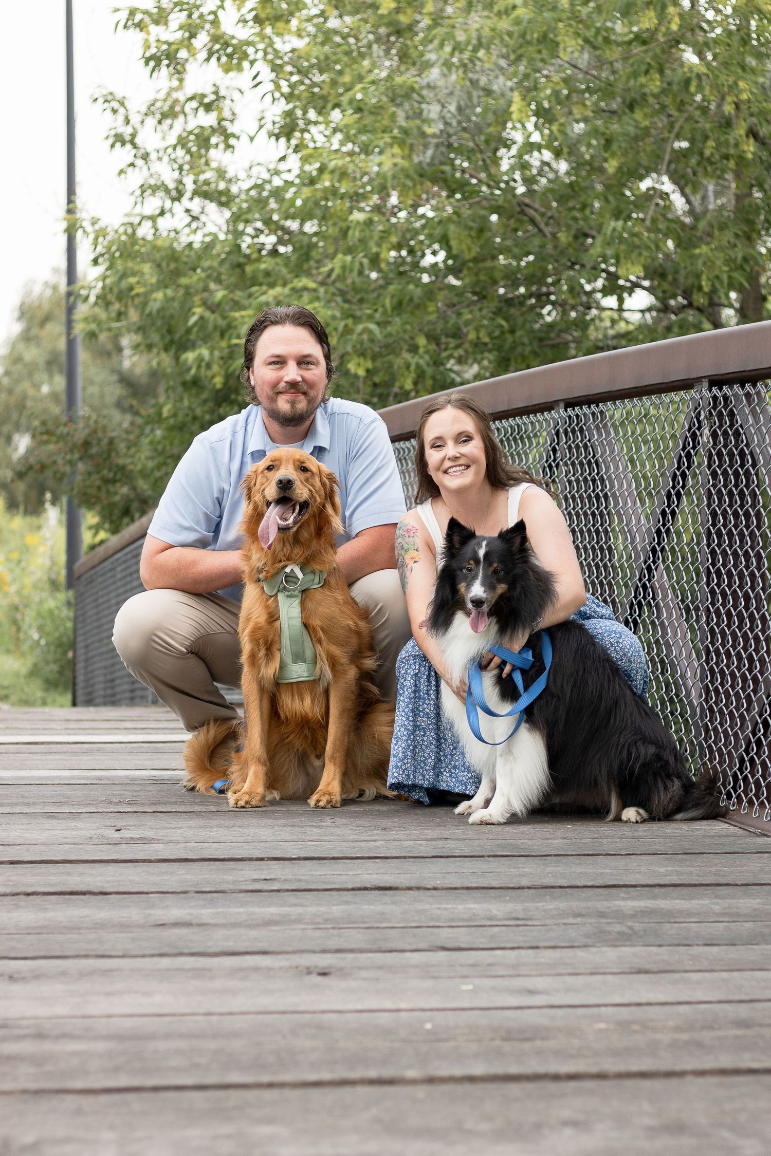 A couple poses with their two dogs on a wooden boardwalk surrounded by greenery.