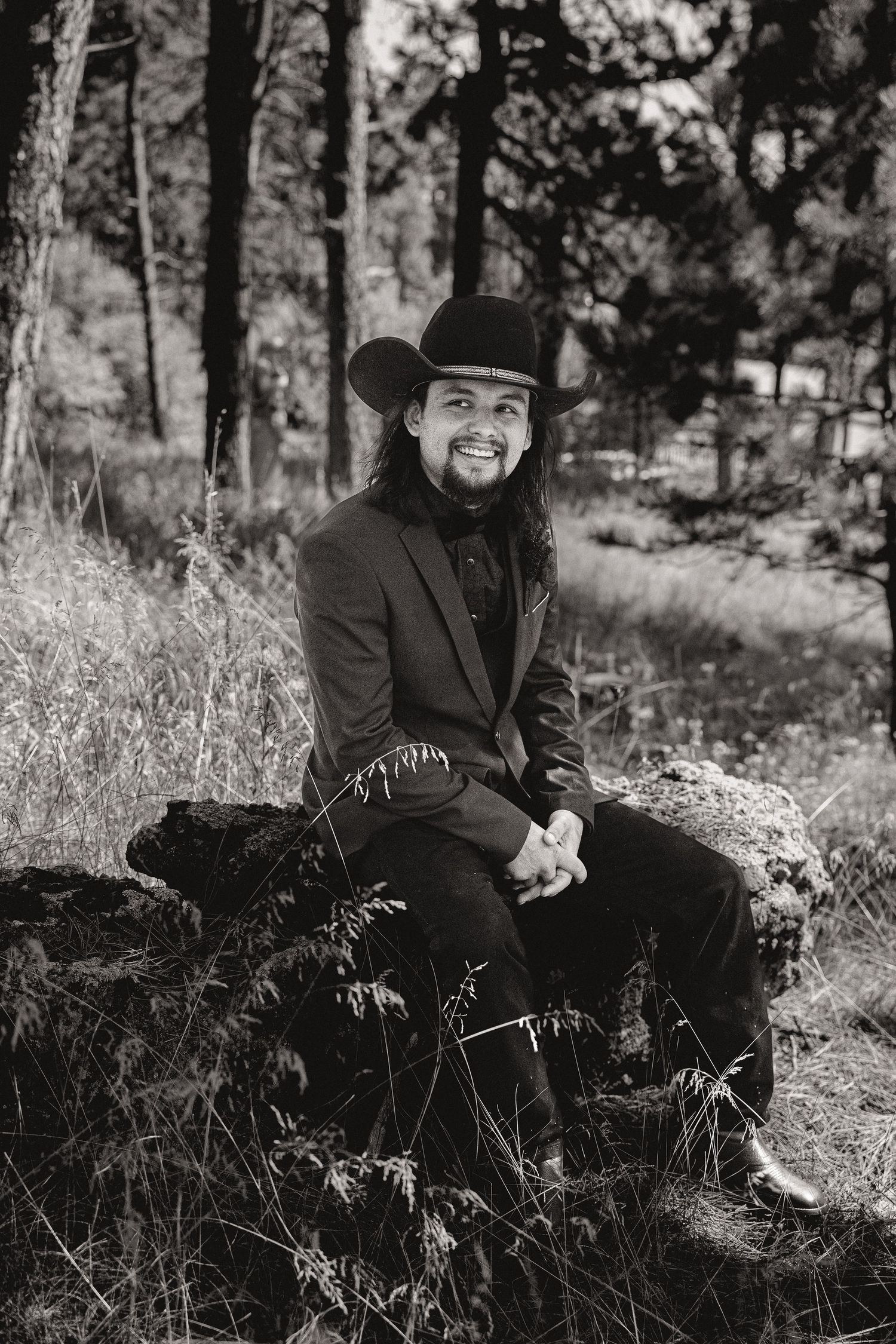 Person in black formal attire and cowboy hat sitting in outdoor setting.
