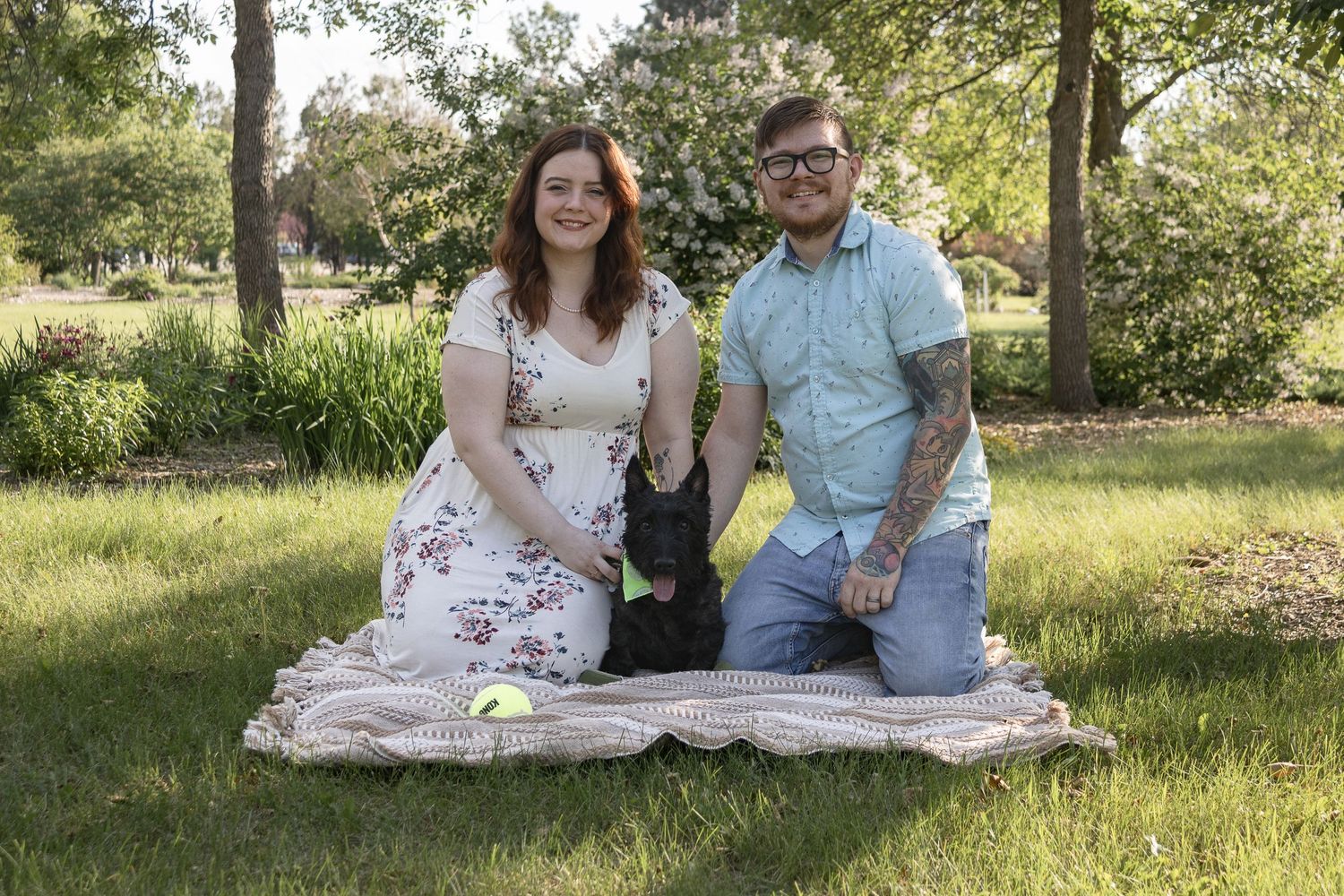 Couple sits on picnic blanket with their black dog in sunny garden setting.