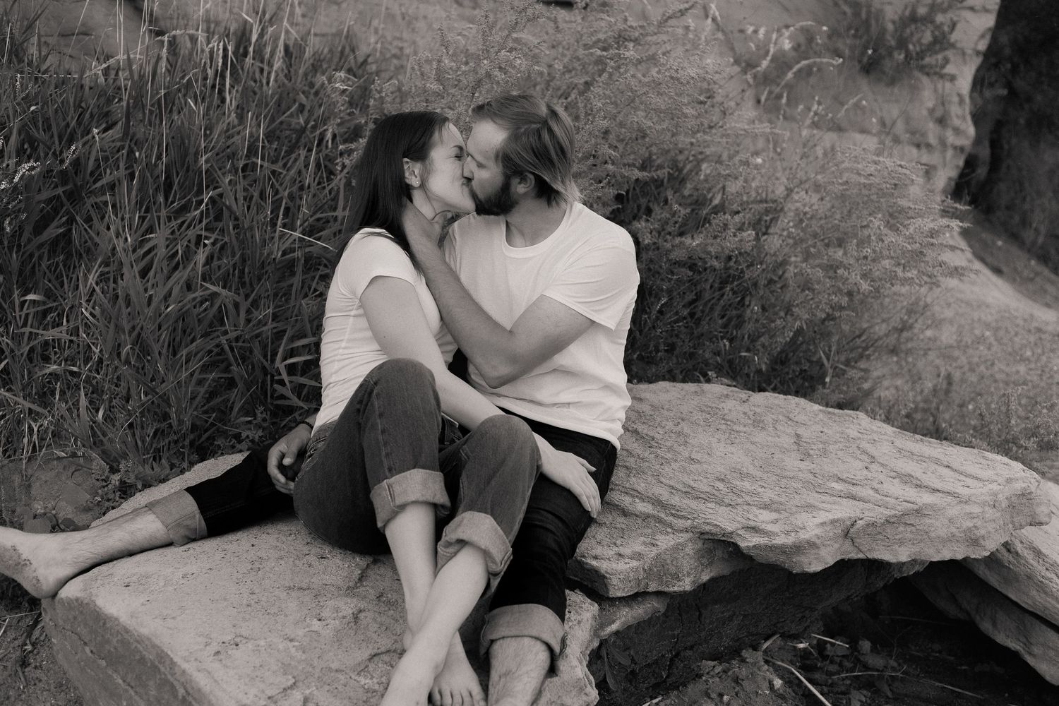 A romantic black and white photo of a couple sitting together on rocks near tall grass.