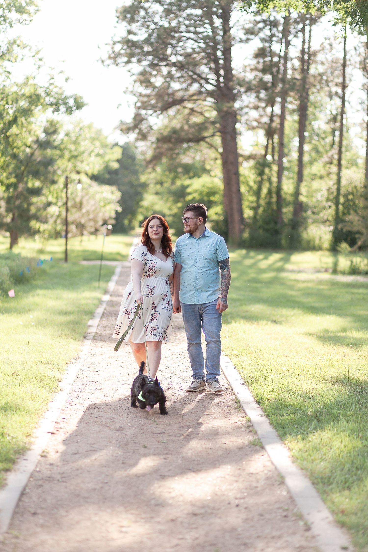 A couple walking their small black dog along a tree-lined path in a park.