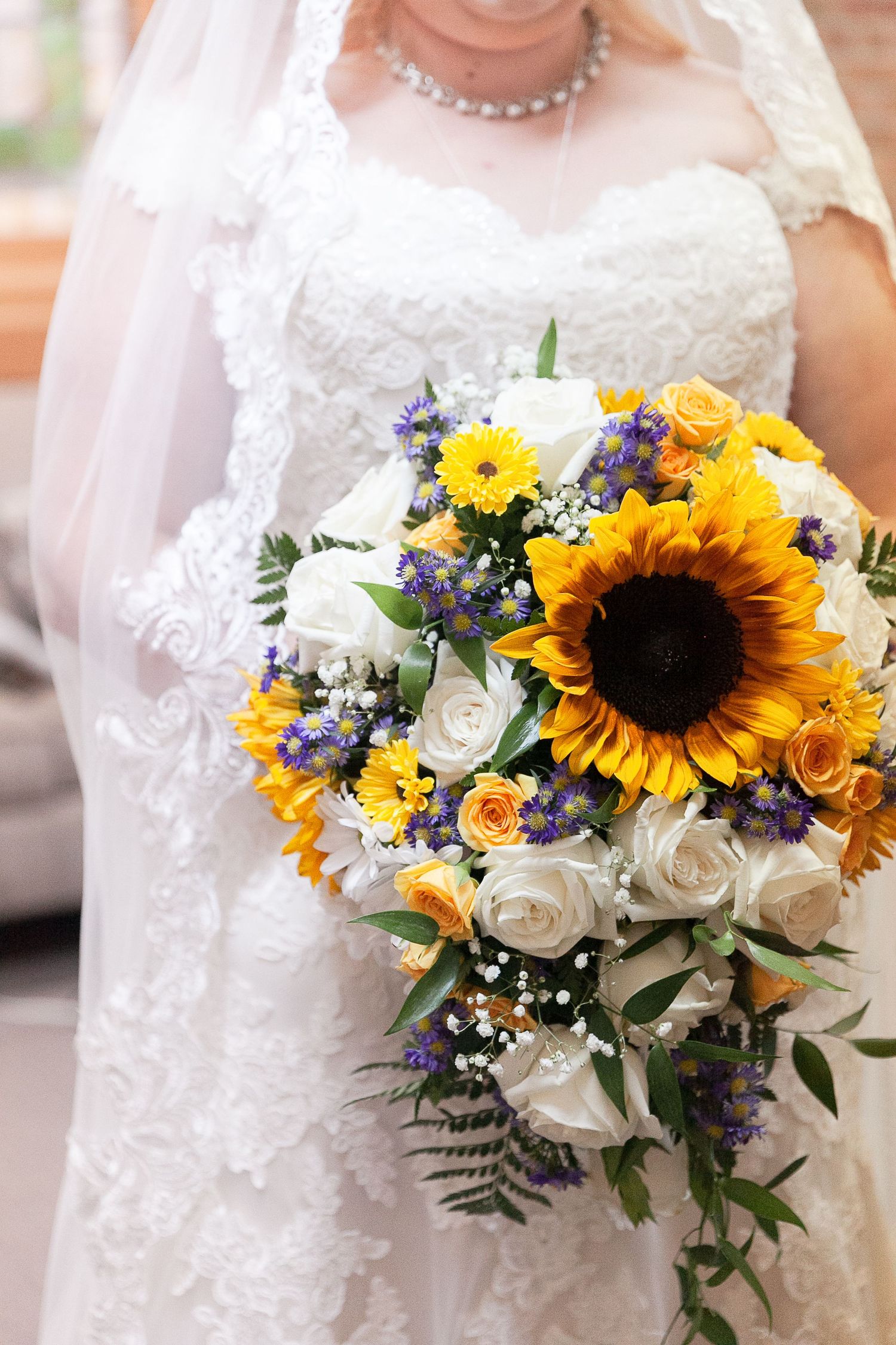 A vibrant bridal bouquet featuring sunflowers, white roses, and purple accents against a lace wedding dress.