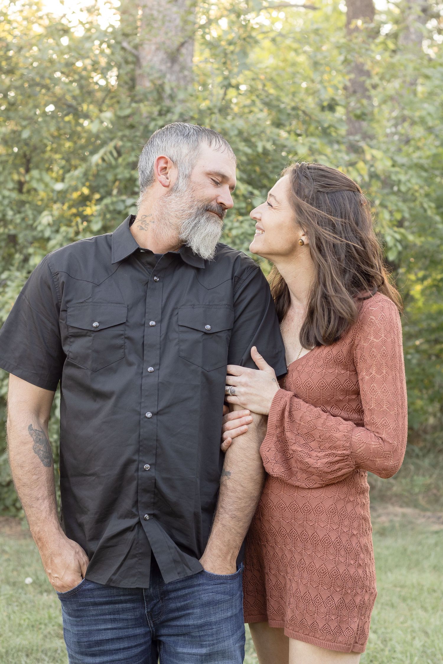 Couple in casual attire share a tender moment while standing together in a wooded outdoor setting.