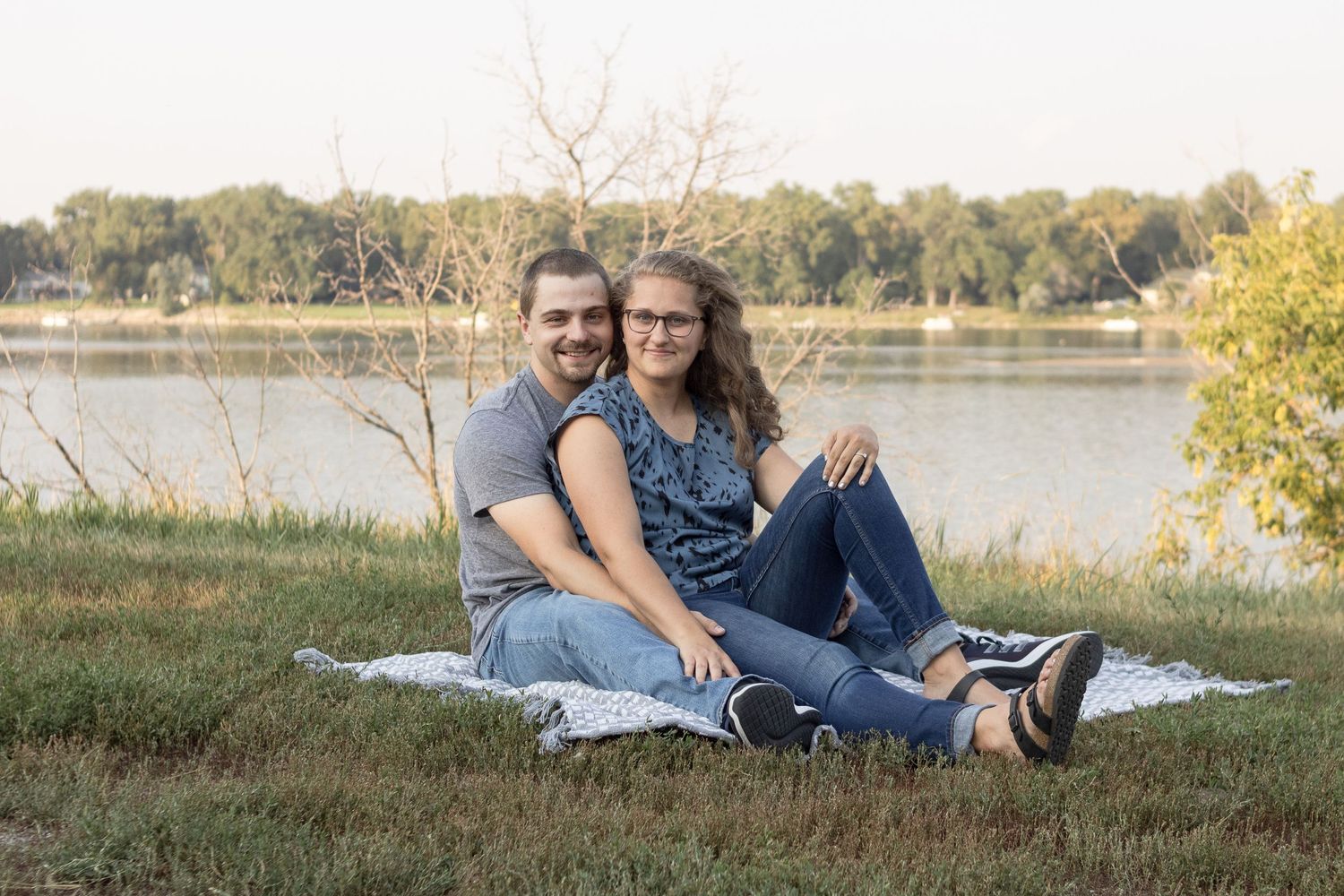 A couple sits together on a blanket by a lake during golden hour.