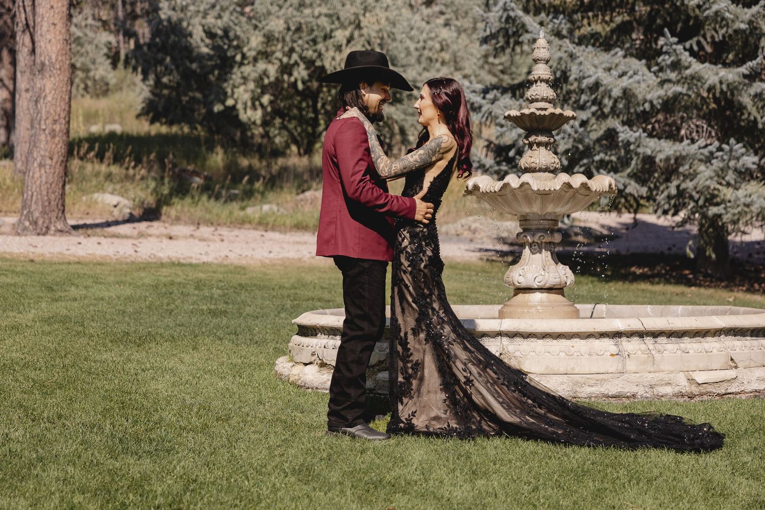 Couple in formal attire embracing outdoors with a long black dress trailing on the grass.