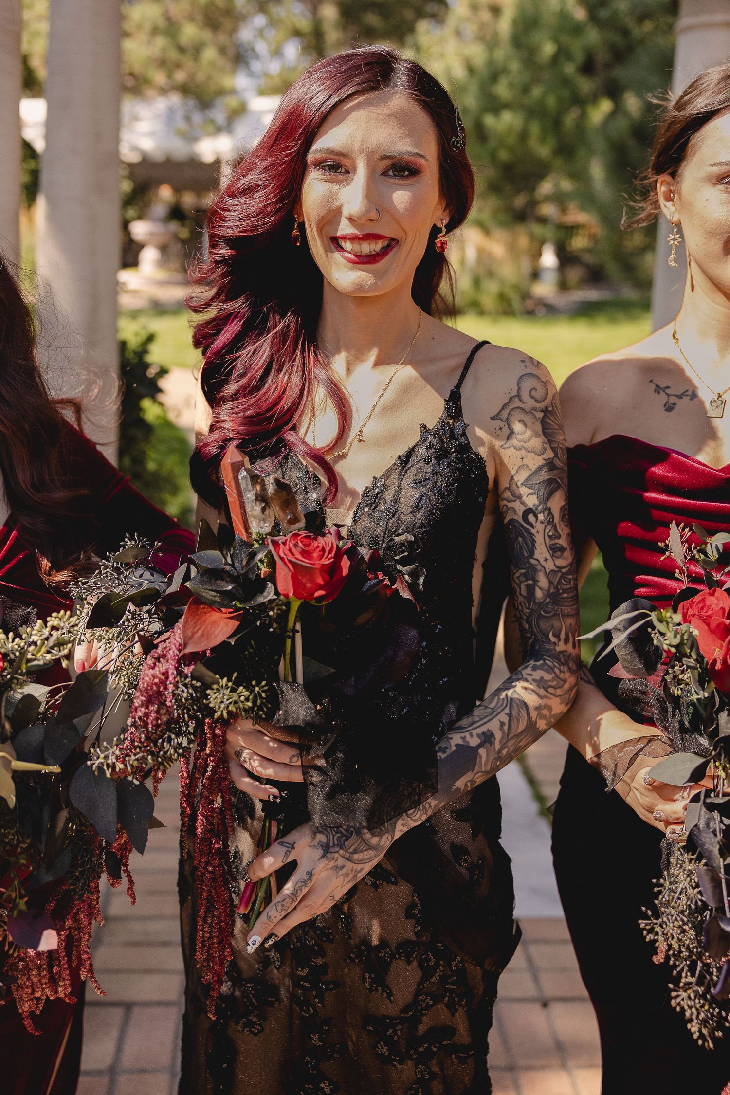 Wedding party member in black lace dress holding red roses at outdoor venue.