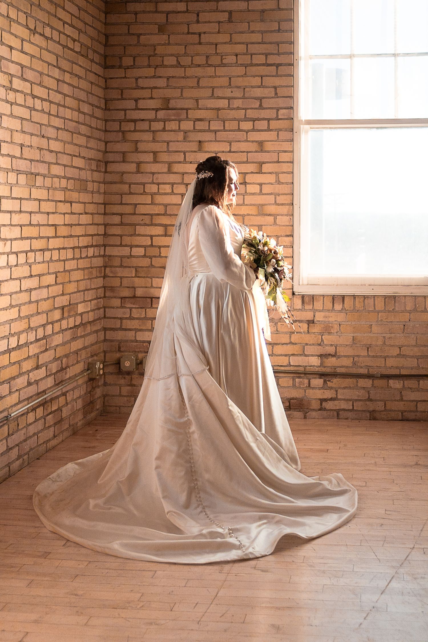 Elegant wedding dress with flowing train photographed against exposed brick wall with natural window light.