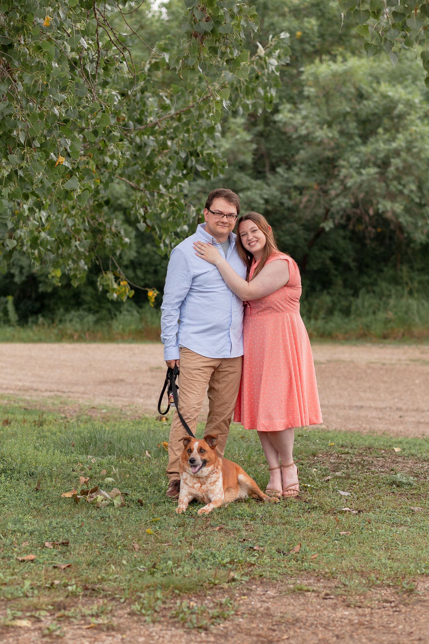 A couple poses with their dog in a park setting, wearing coordinated casual outfits.